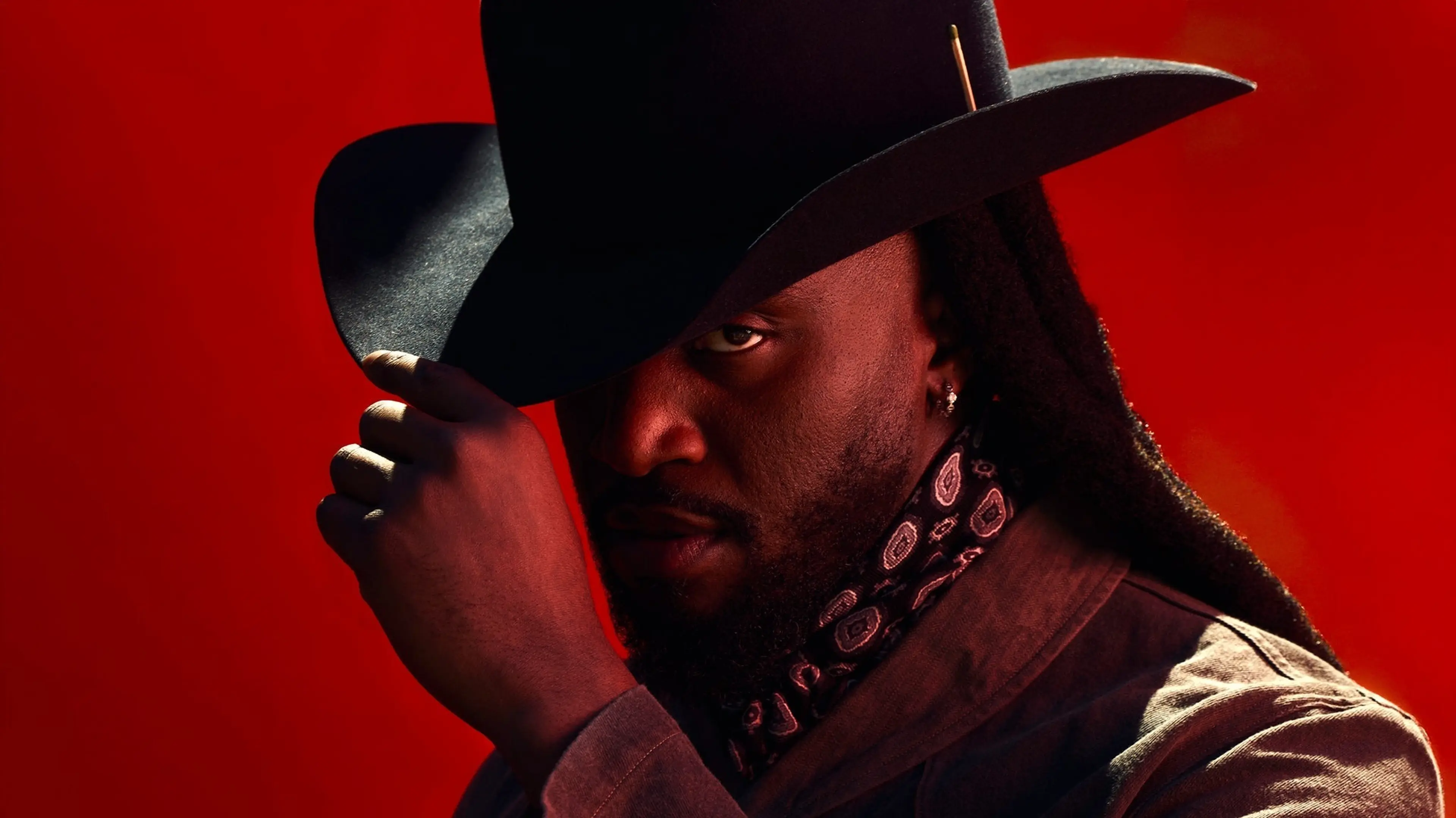 Person wearing a black wide-brimmed hat and patterned scarf against a red background, partially covering their face with the hat.