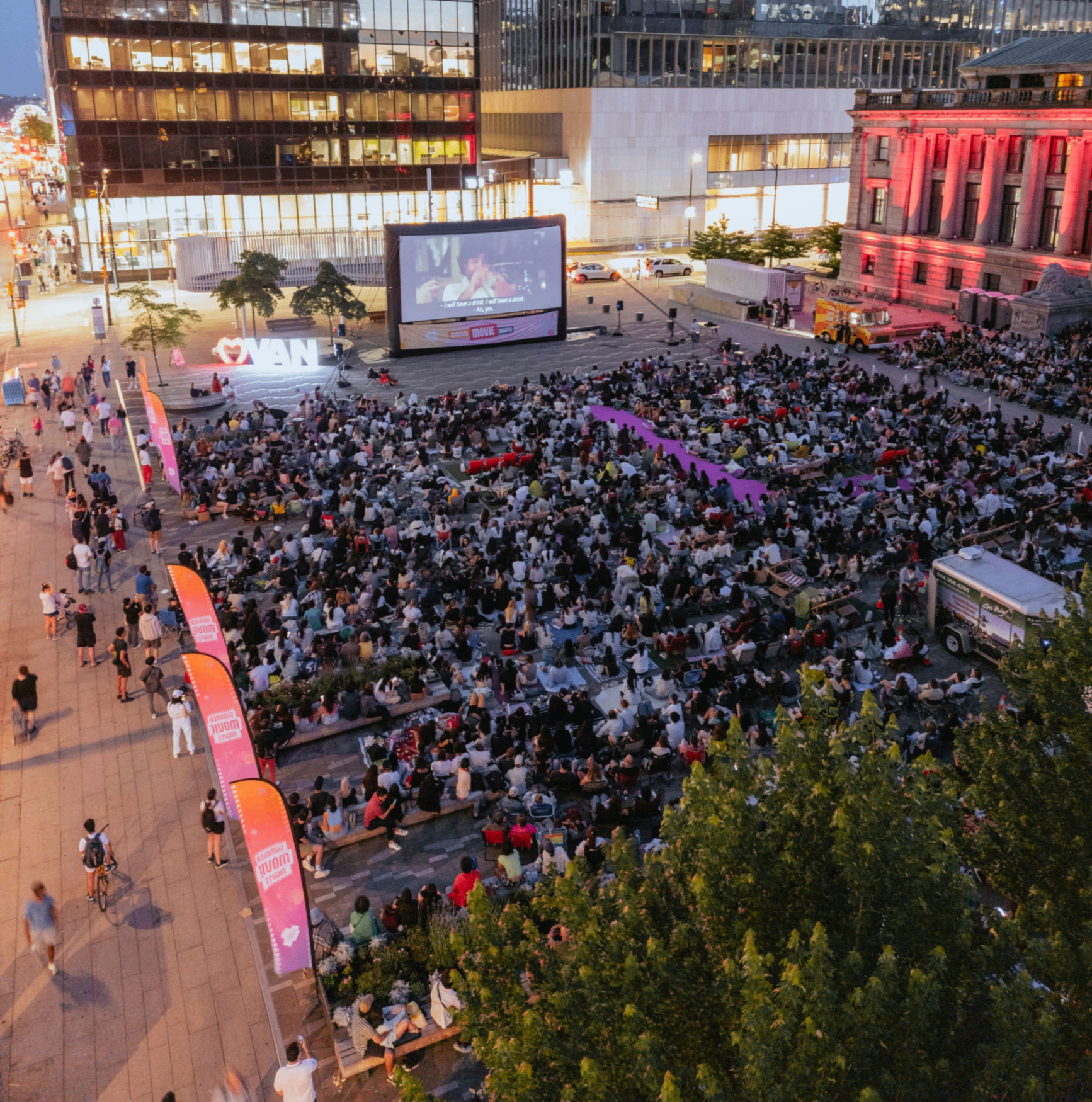 Large crowd watching an outdoor movie screening in a city plaza at dusk with illuminated buildings and trees nearby.