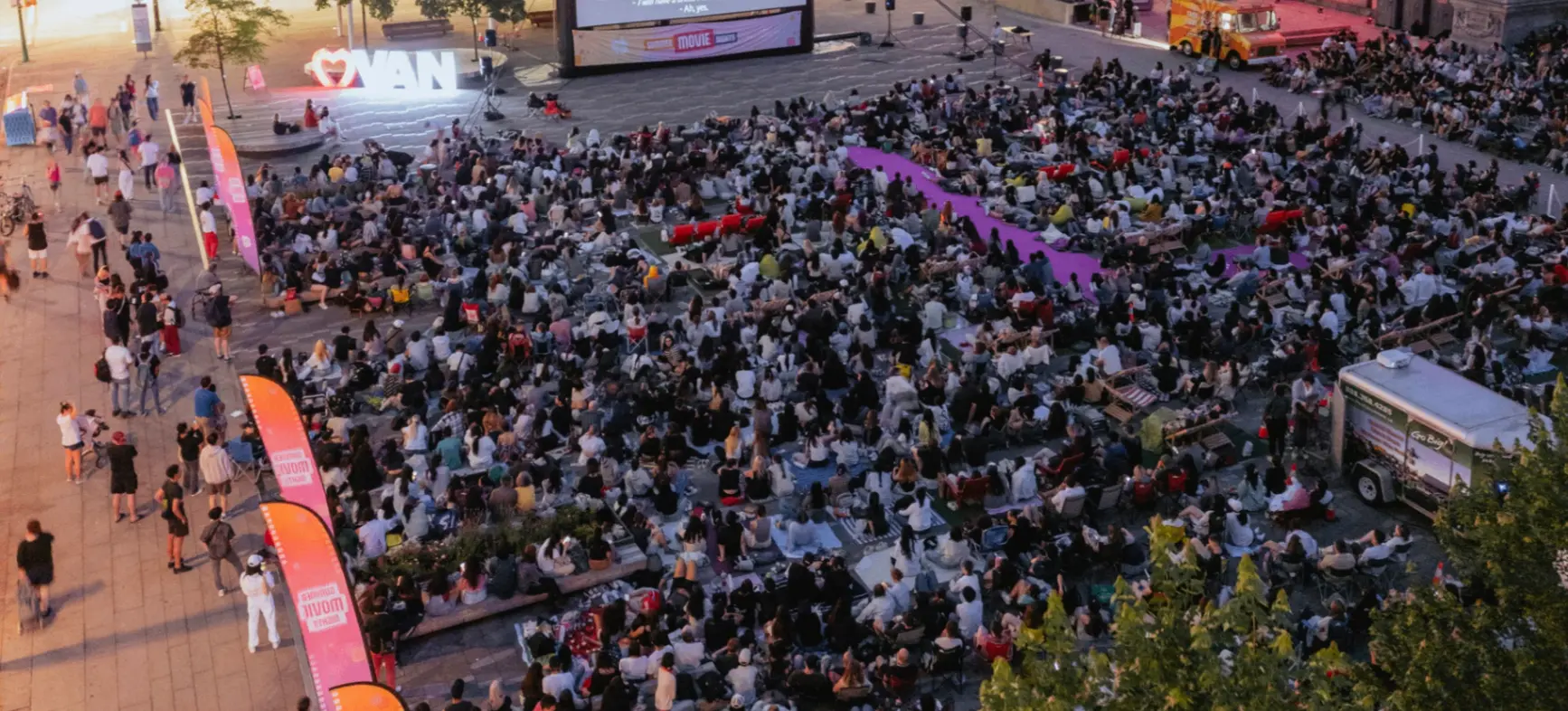 Large crowd watching an outdoor movie screening in a city plaza at dusk with illuminated buildings and trees nearby.