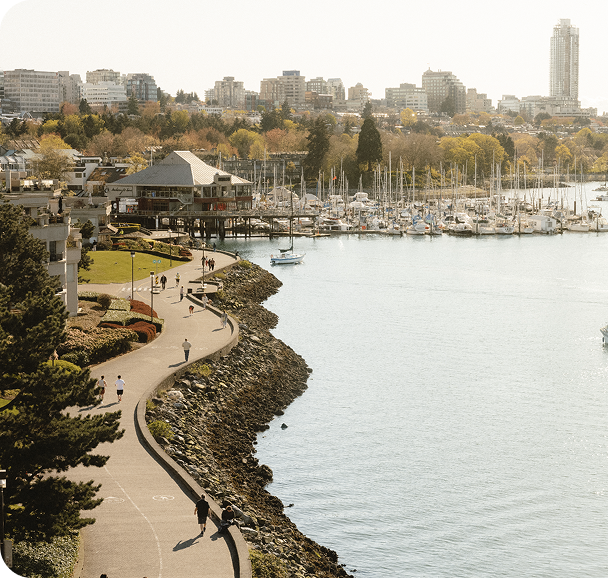 Curved waterfront path with people walking and jogging, marina with boats, and city buildings in the background.