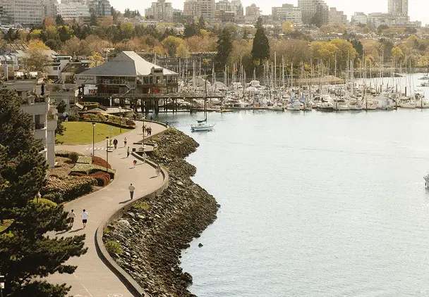 Curved waterfront path with people walking and jogging, marina with boats, and city buildings in the background.