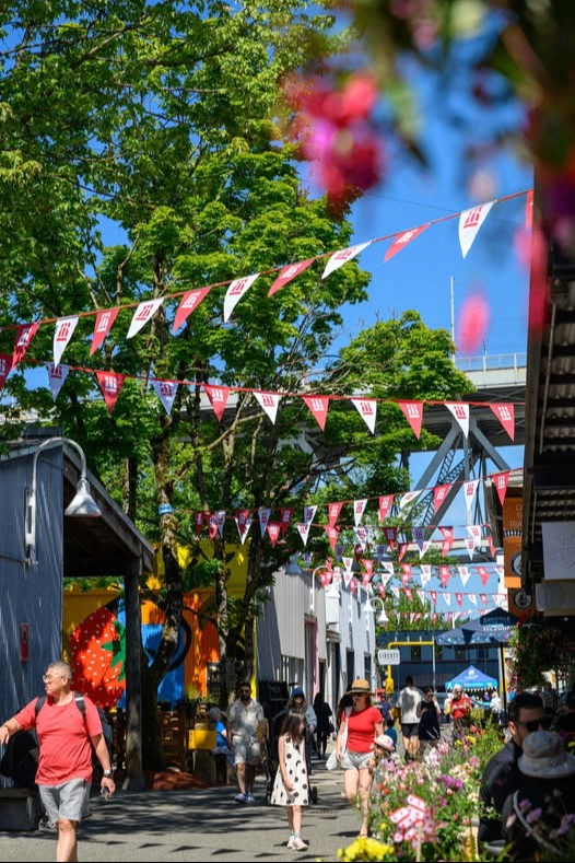 Outdoor street scene with people walking under red and white triangular flags, green trees, and colorful flowers on a sunny day.