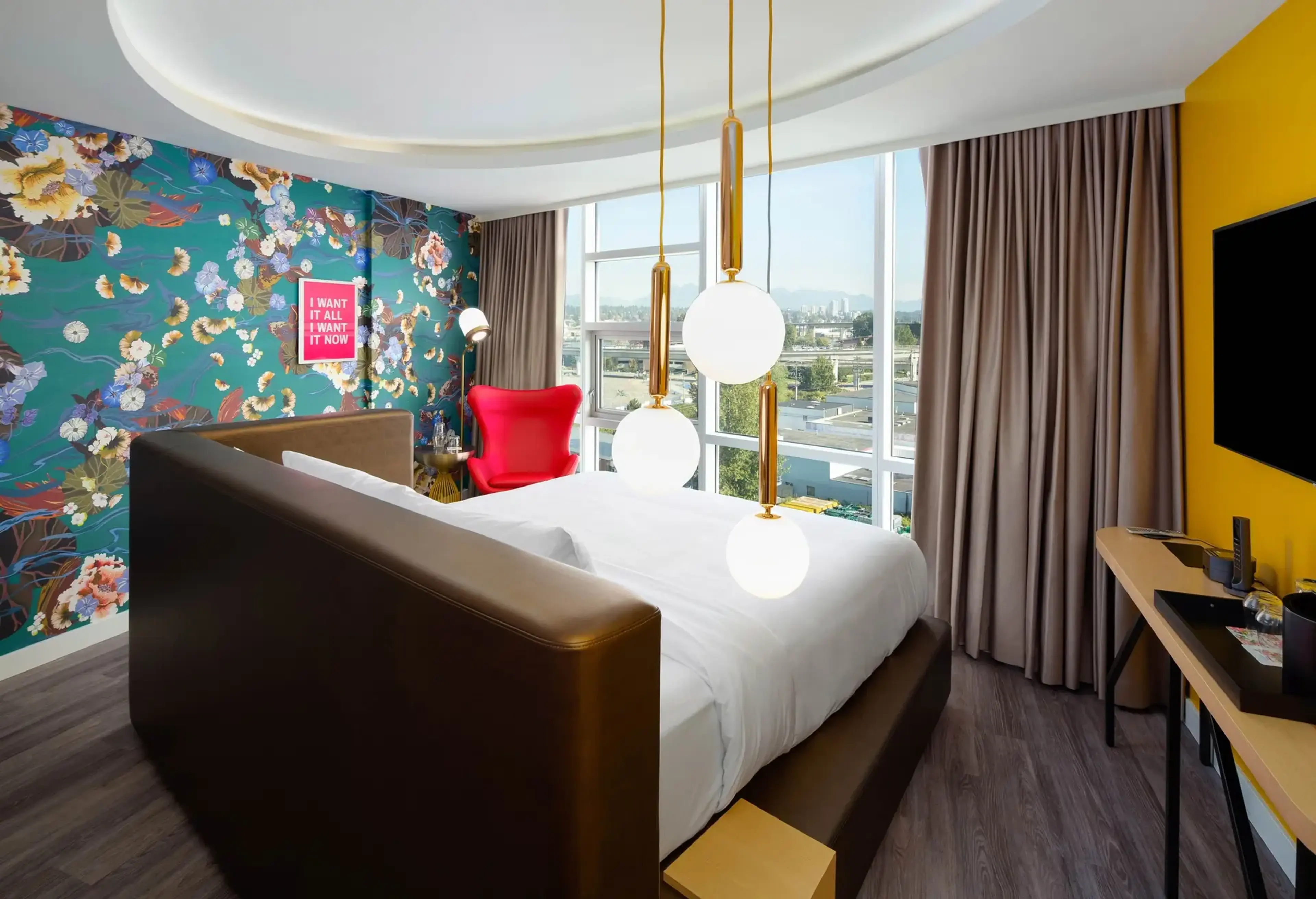 Modern bedroom with floral wallpaper, large bed, red armchair, hanging globe lights, and city view through large windows.