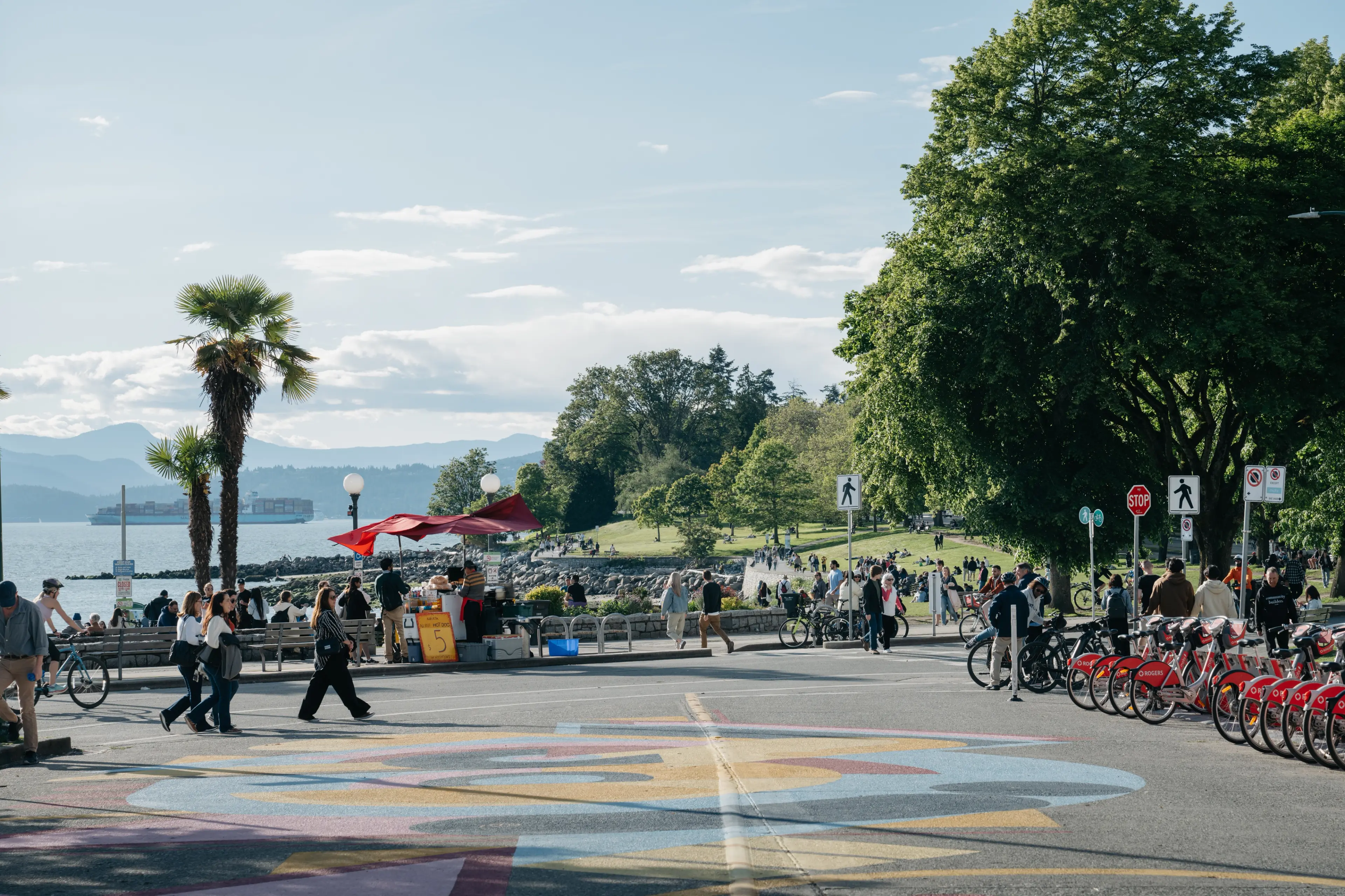 People walking and biking near a waterfront park with trees, a food stand with a red umbrella, and a row of red rental bikes.