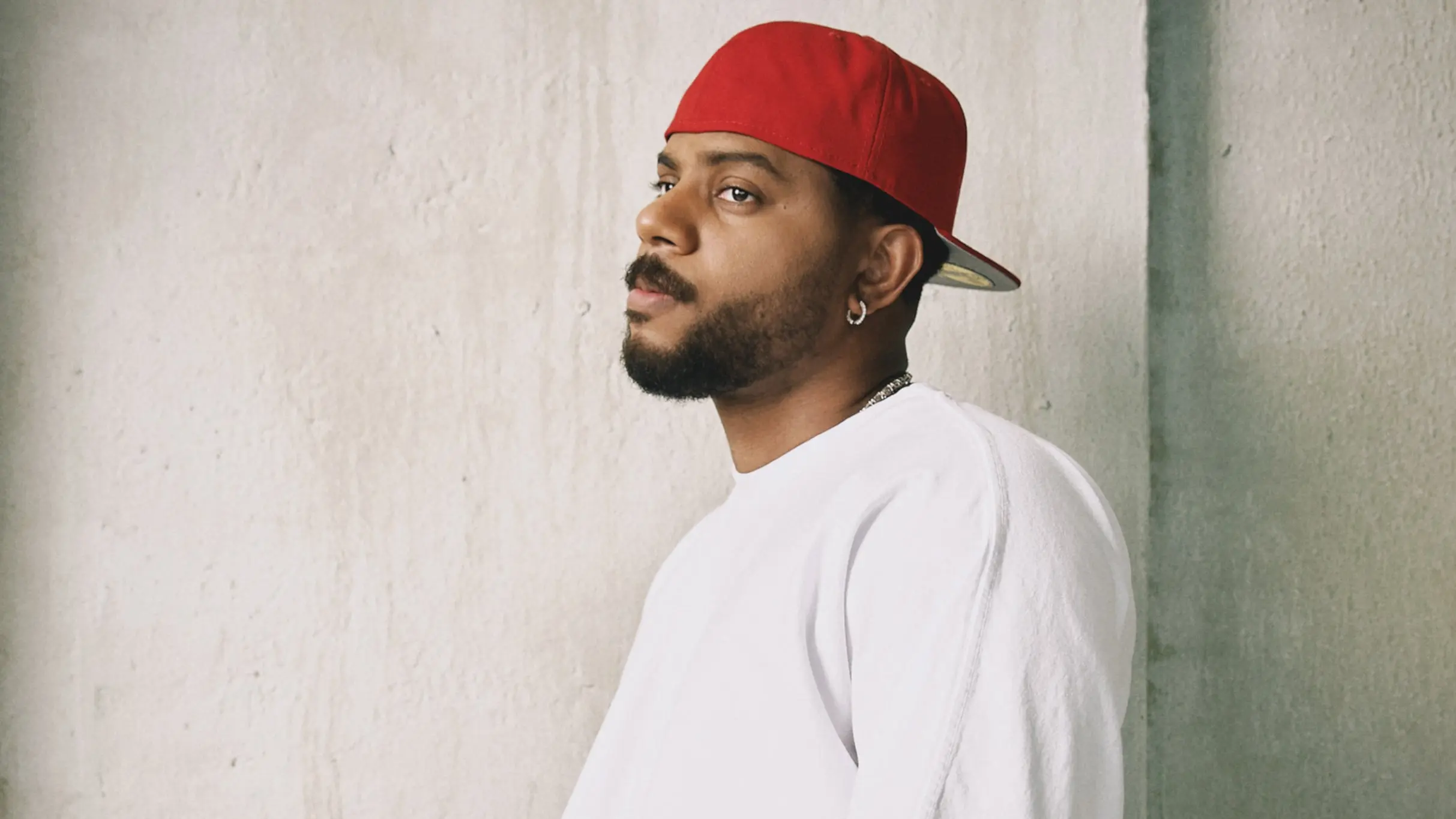 Person wearing a red backward cap, white shirt, and hoop earring looking to the side against a plain wall.