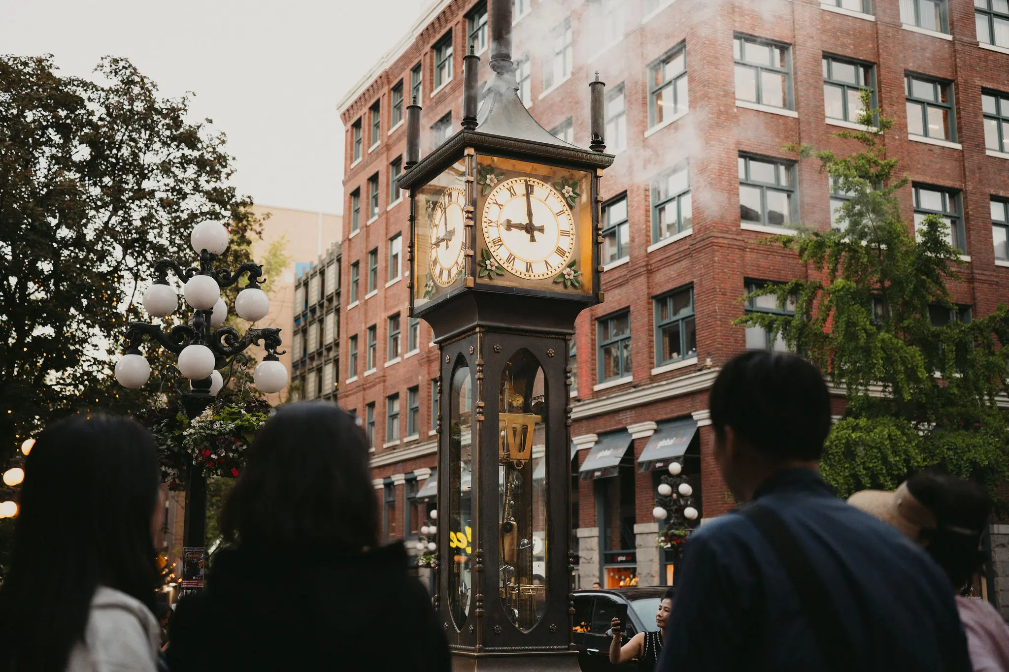Steam clock on a city street with people looking at it and a brick building in the background.