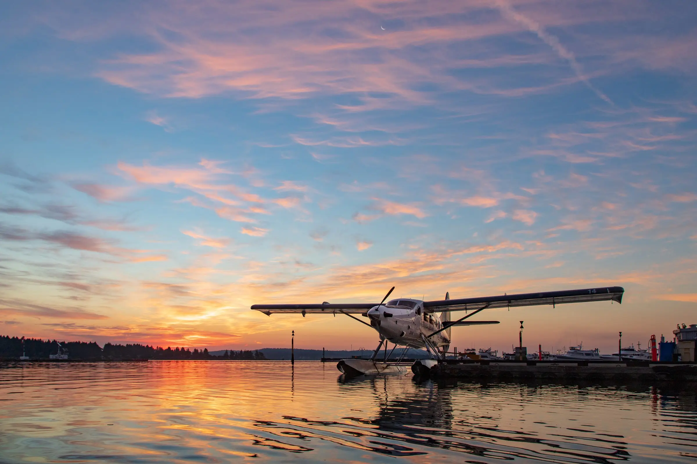A floatplane rests on calm water at sunset, with colorful clouds and a vibrant sky reflecting on the surface—a scene straight from a Vancouver travel guide. Distant boats and a shoreline are visible in the background.