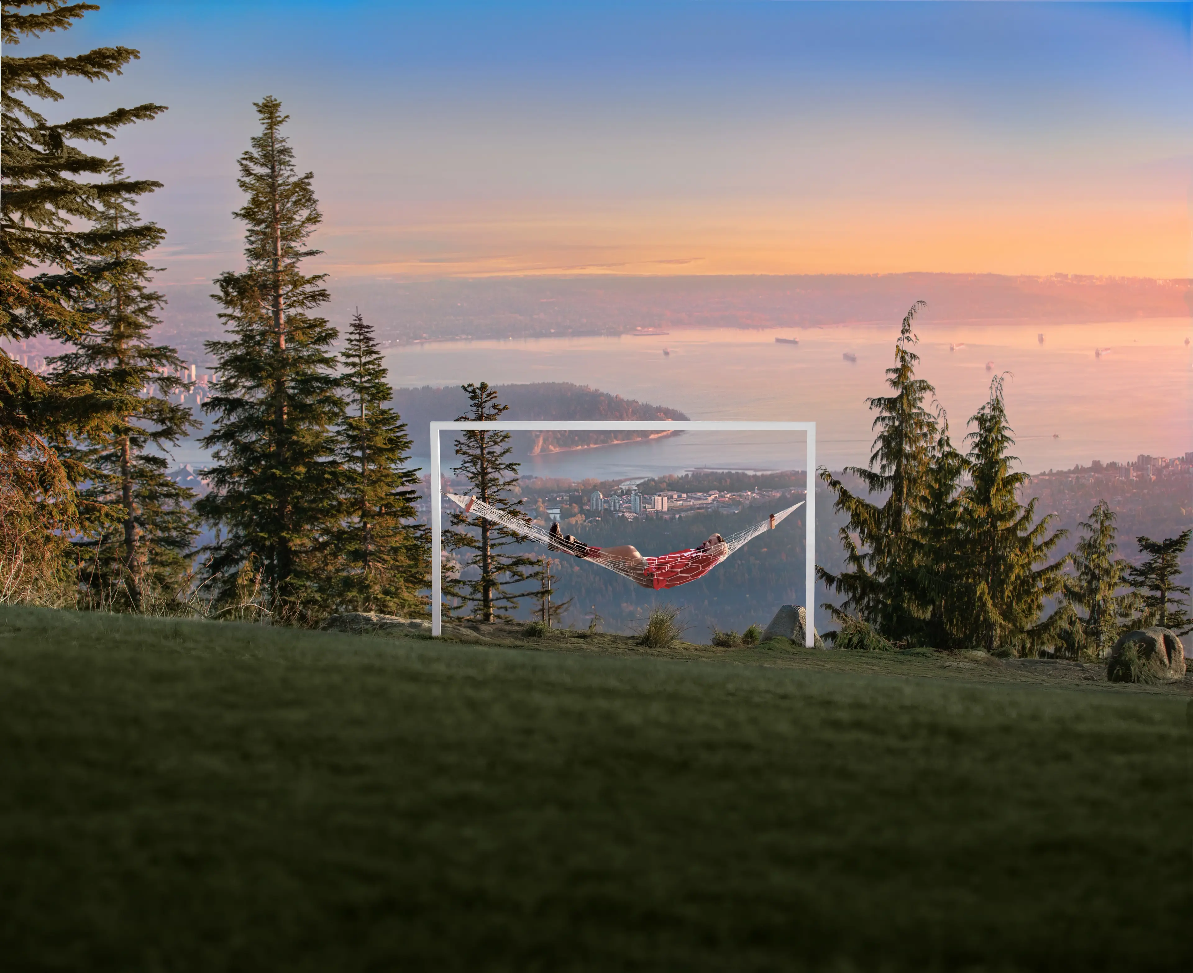 Person lying in a hammock suspended on a white frame on a grassy hill with trees and a city and water view at sunset.