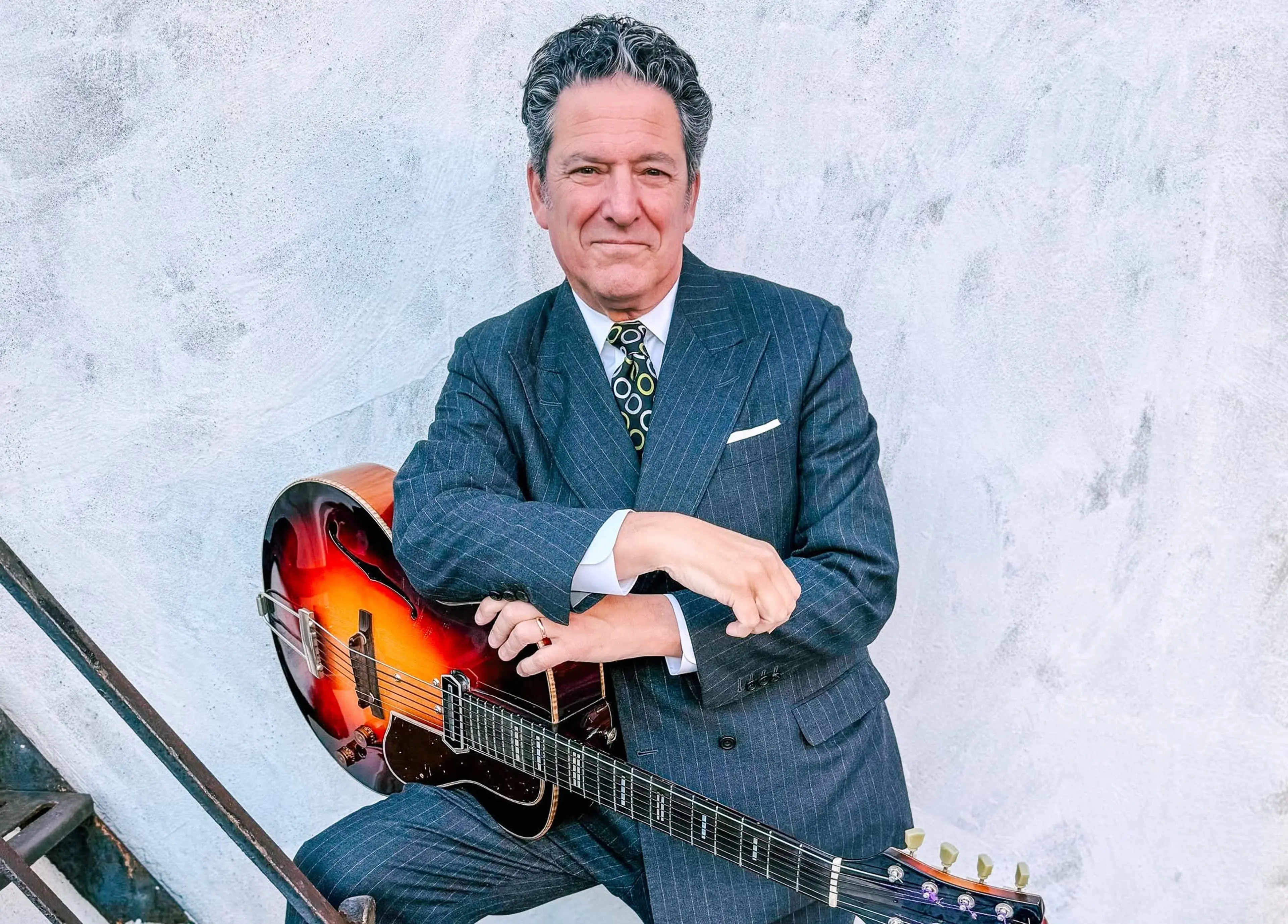 Person in a pinstripe suit sitting with a sunburst electric guitar against a textured light wall.