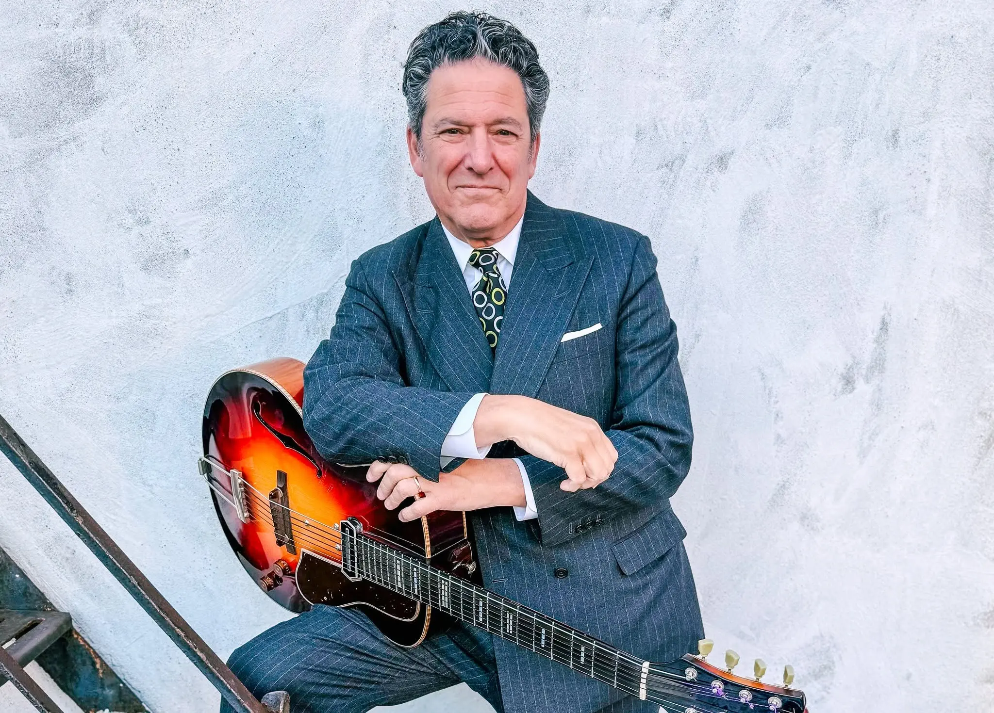 Person in a pinstripe suit sitting with a sunburst electric guitar against a textured light wall.