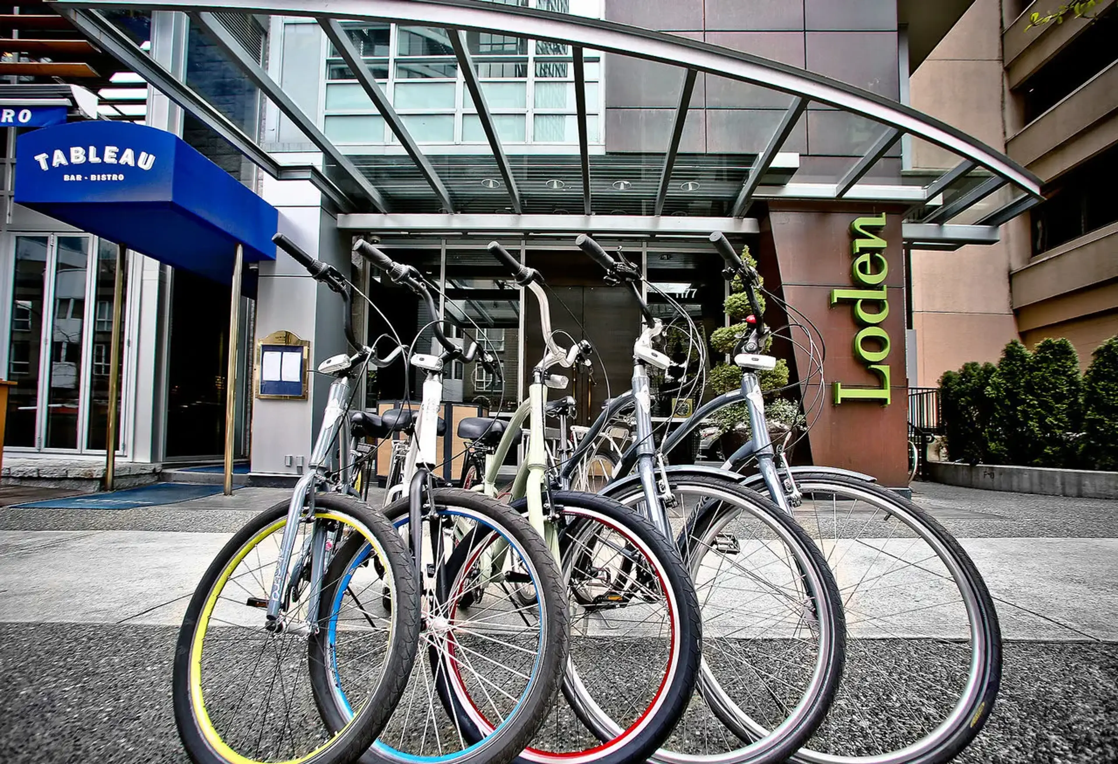 Five bicycles parked in front of a building with a glass canopy and signs reading Tableau Bar Bistro and Lodlen.