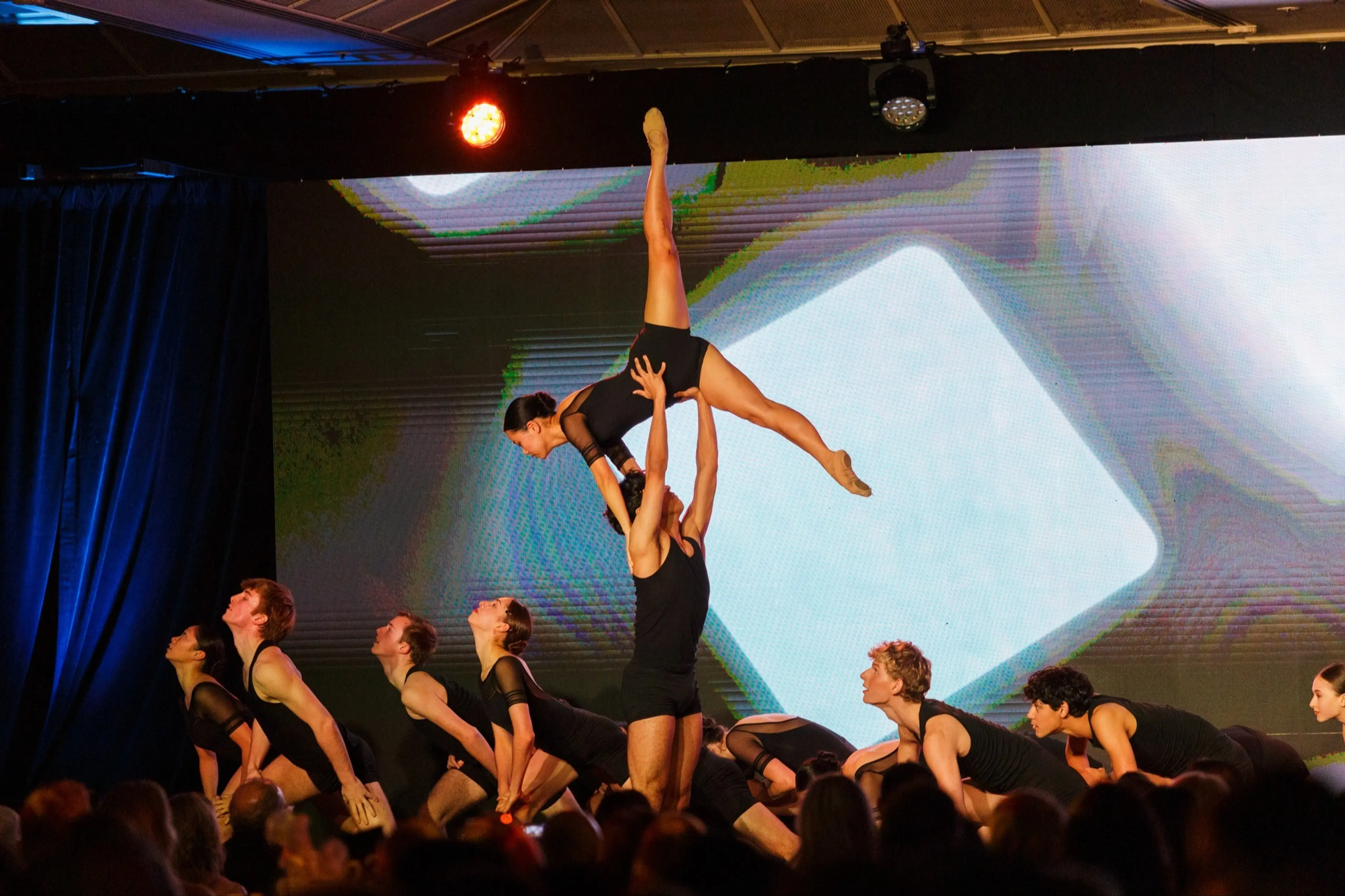 A group of dancers in black outfits performing on stage, one dancer is lifted high with legs extended vertically.