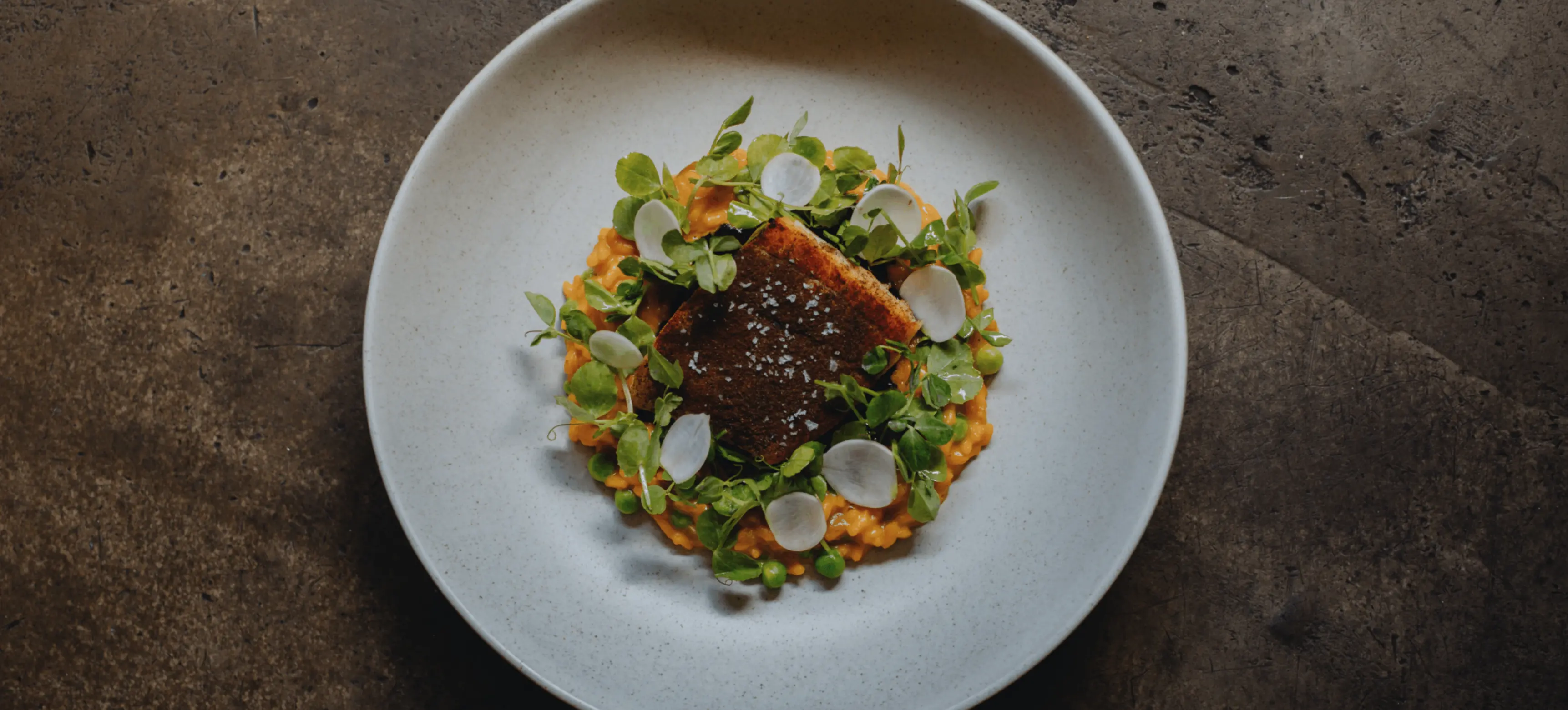 Grilled fish fillet on orange puree, garnished with green leaves and white radish slices on a white plate.