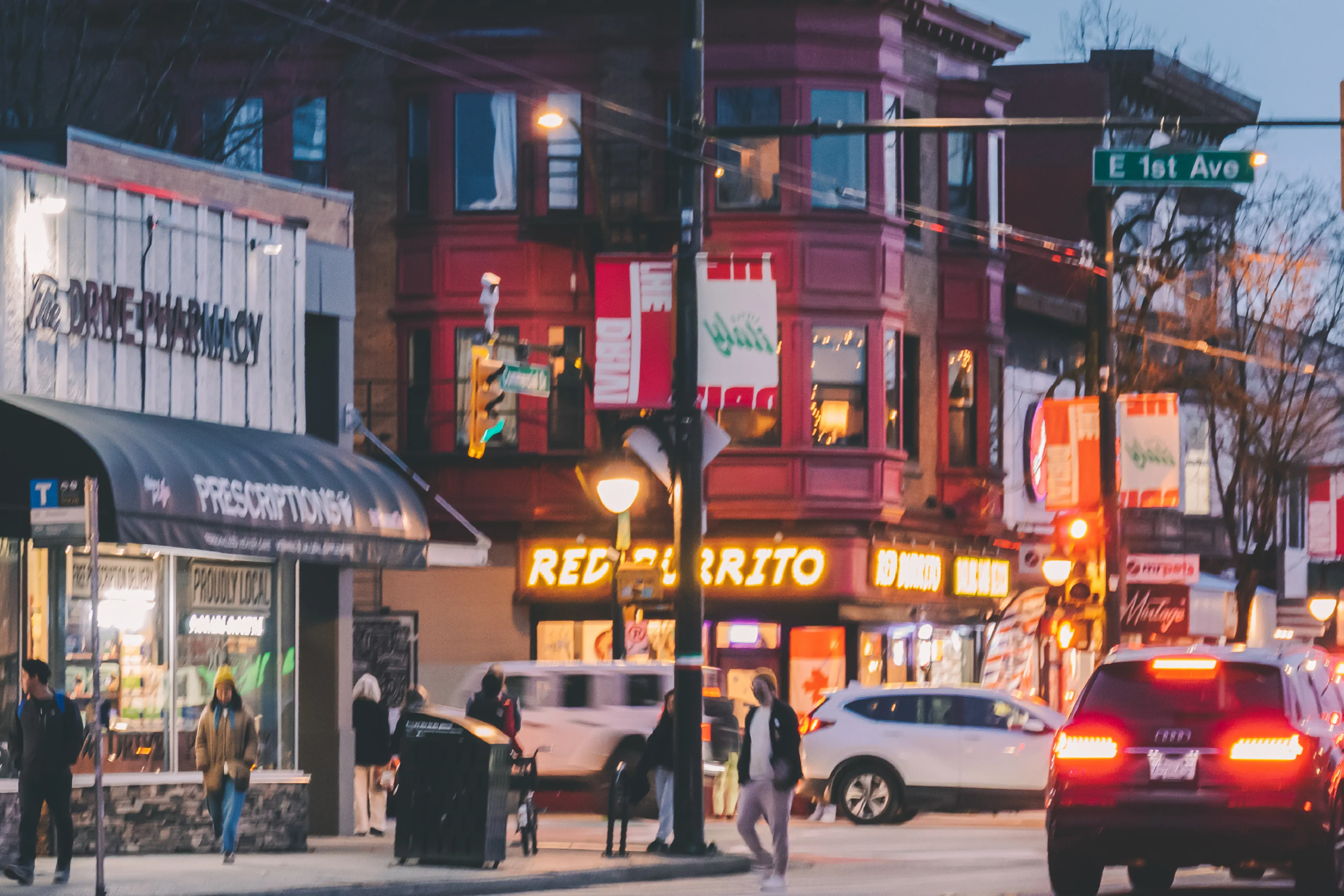 Street scene at dusk with pedestrians, cars, a pharmacy on the left, and a red building with a bright 'Red Burrito' sign.