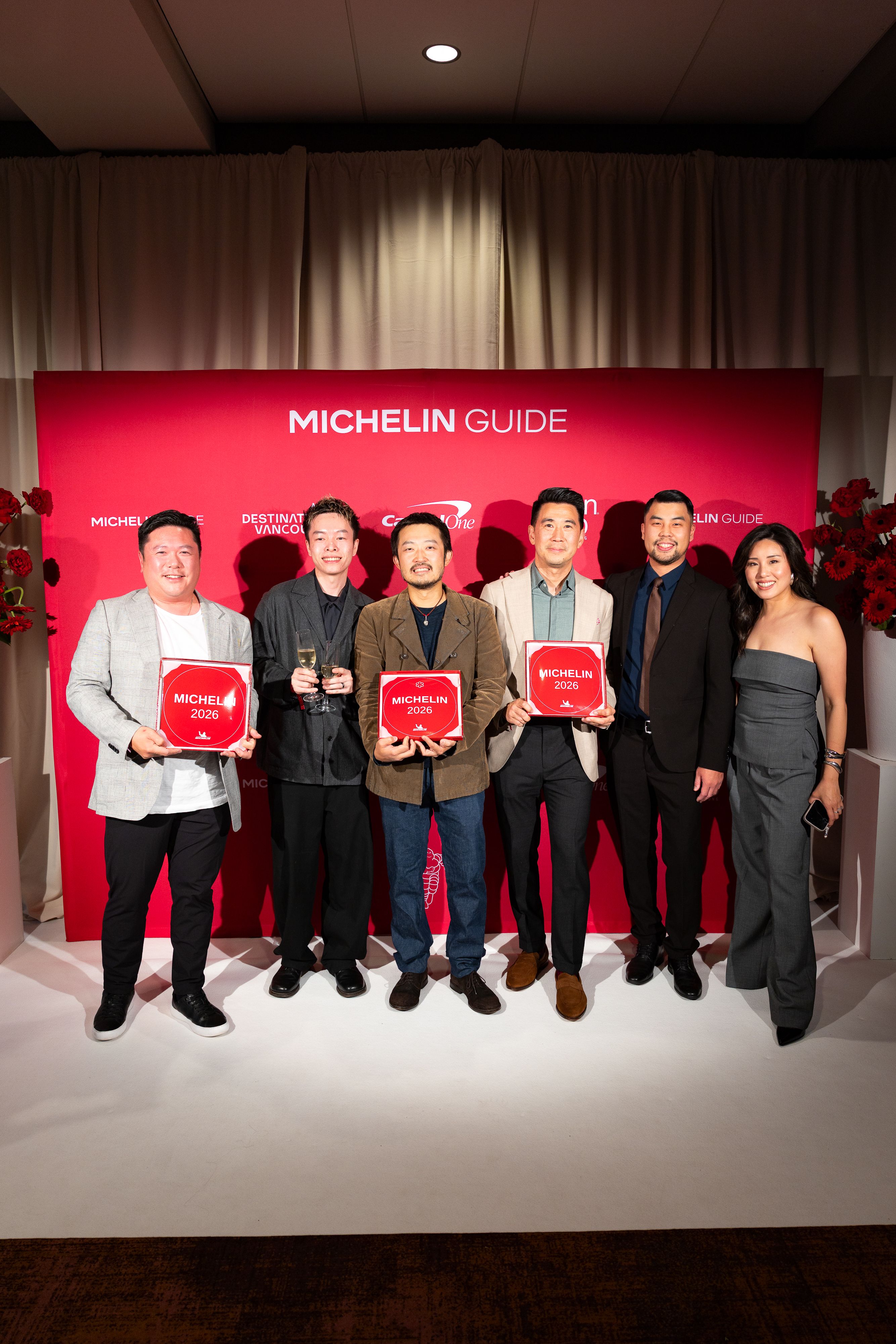 Six people standing in front of a red Michelin Guide backdrop, three holding Michelin 2026 plaques, one holding two glasses of champagne.
