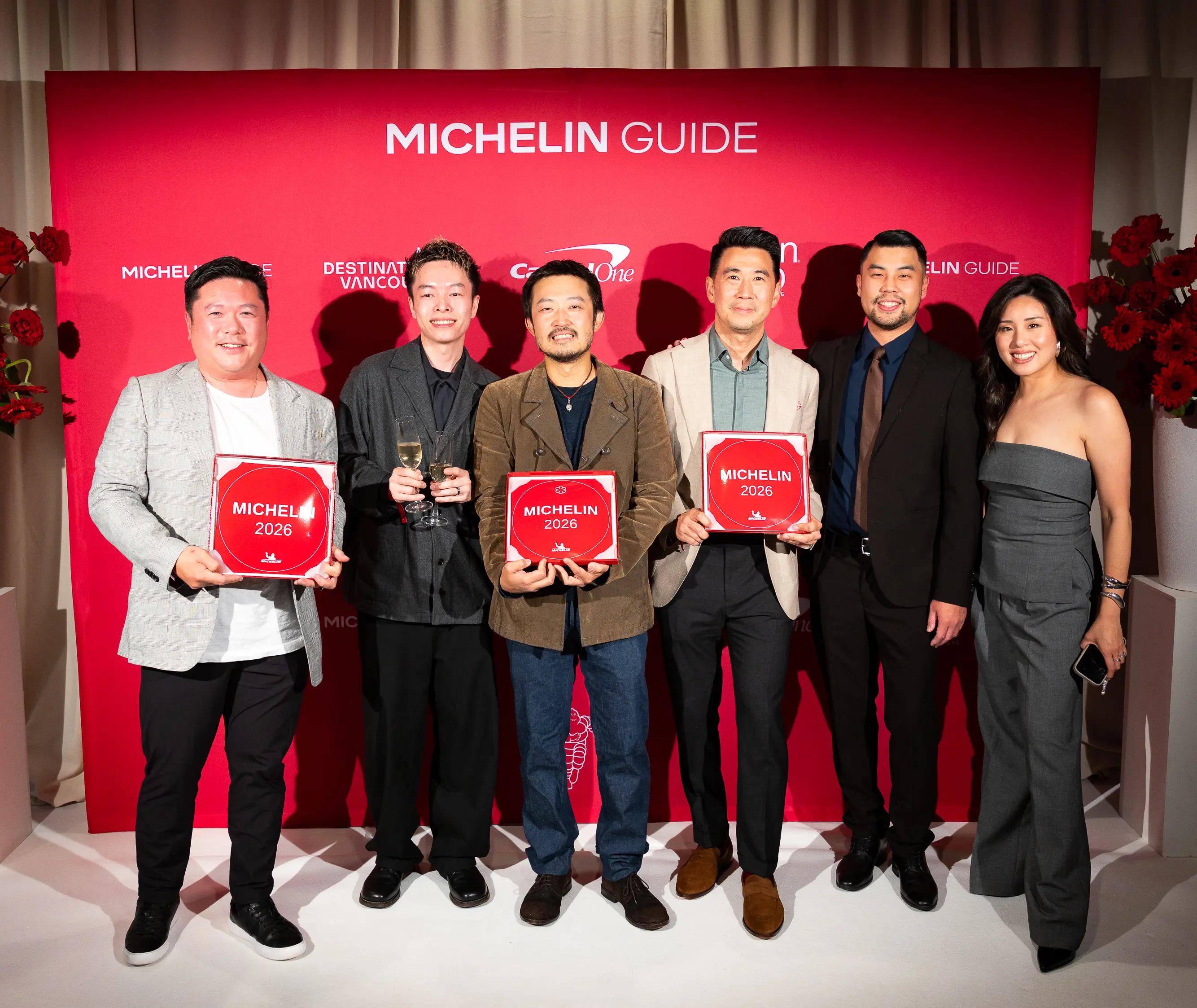 Six people standing in front of a red Michelin Guide backdrop, three holding Michelin 2026 plaques, one holding two glasses of champagne.
