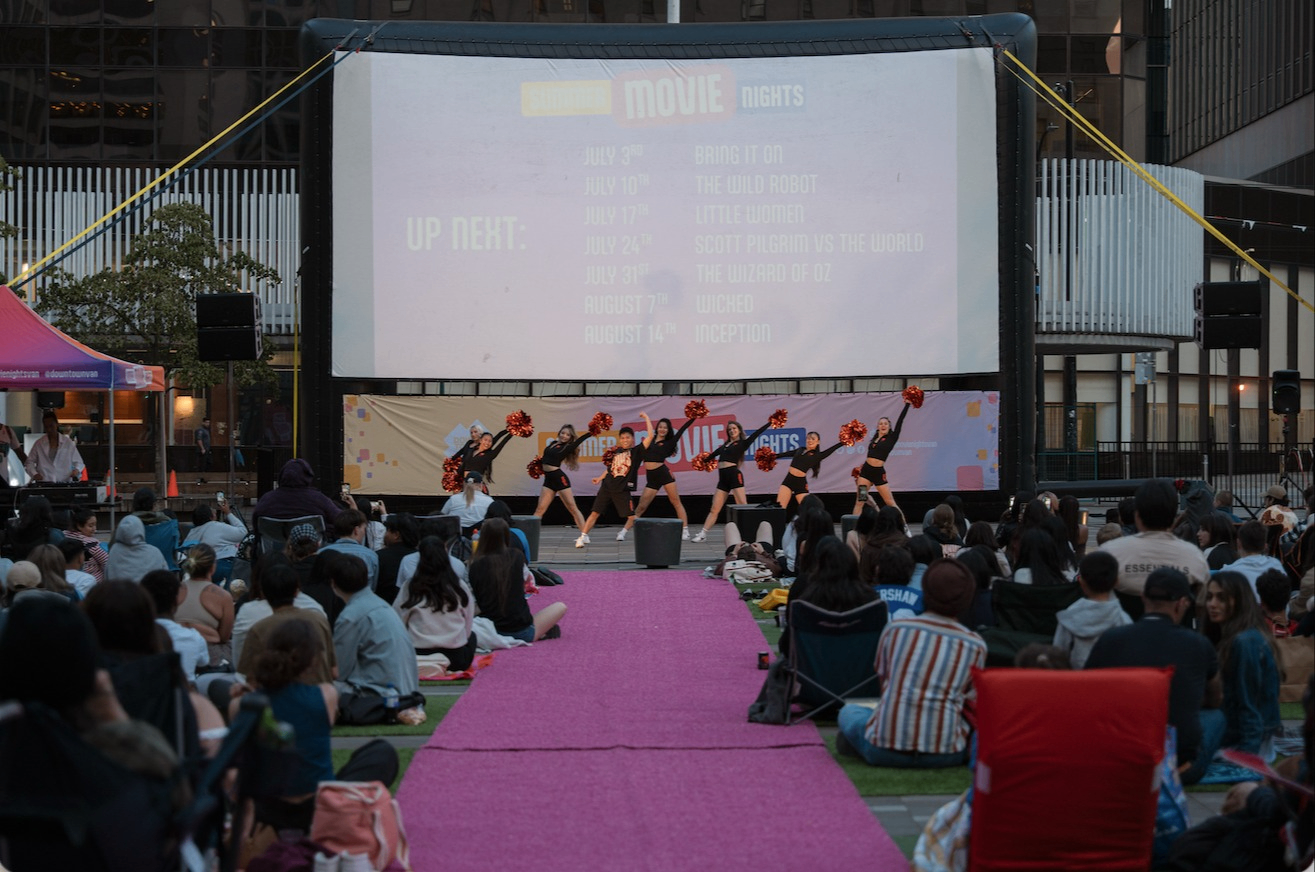 Cheerleaders performing with pom-poms on stage in front of a large outdoor movie screen and seated audience.
