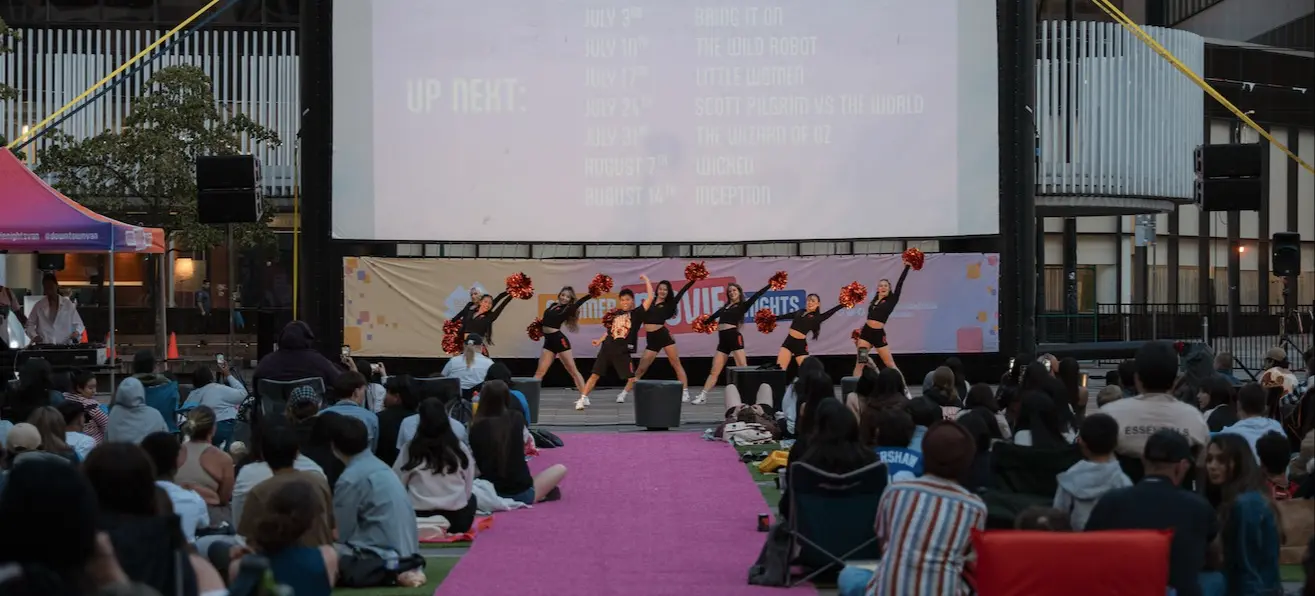 Cheerleaders performing with pom-poms on stage in front of a large outdoor movie screen and seated audience.