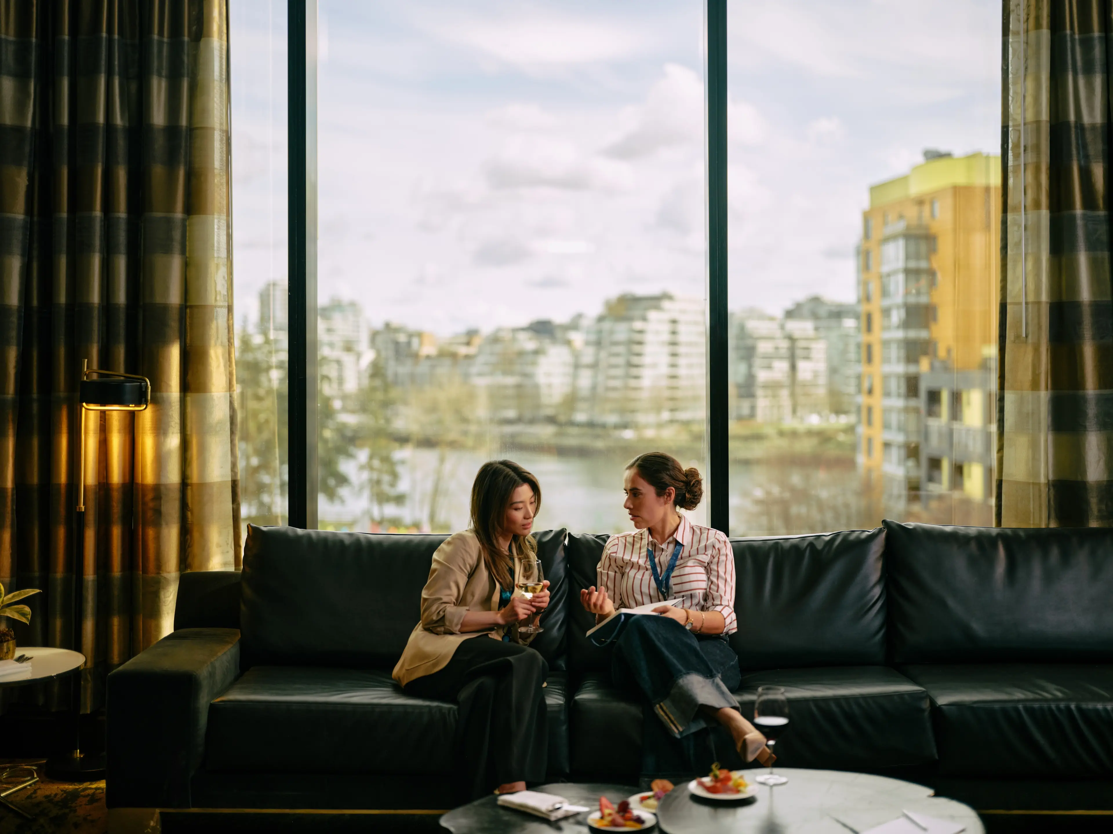 Two delegates sitting on a couch in a hotel room at the JW Marriott Parq Hotel in Vancouver, with False Creek visible through the window in the background.
