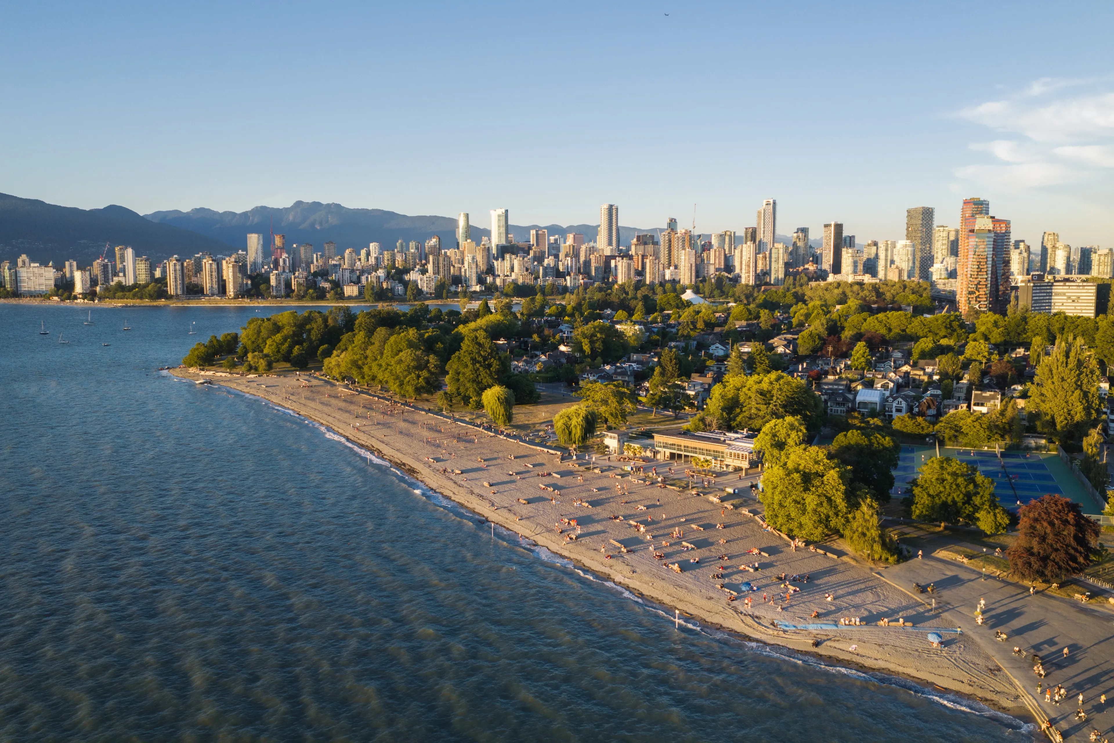 Aerial view of a sandy beach with people, green trees, and a city skyline with tall buildings in the background under a clear sky.