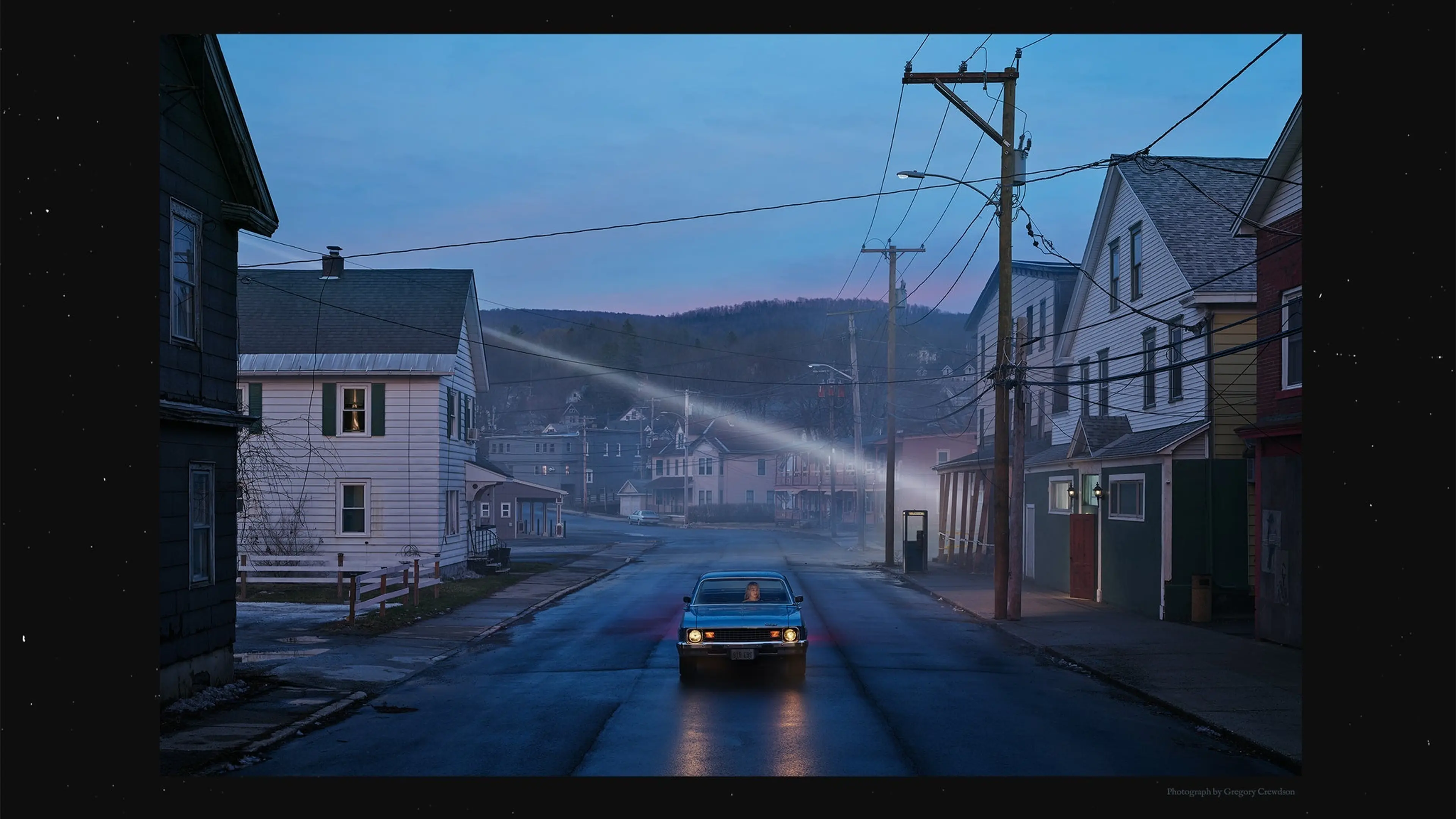 A car with headlights on drives down a quiet street at dusk in a small town with houses and utility poles on both sides.