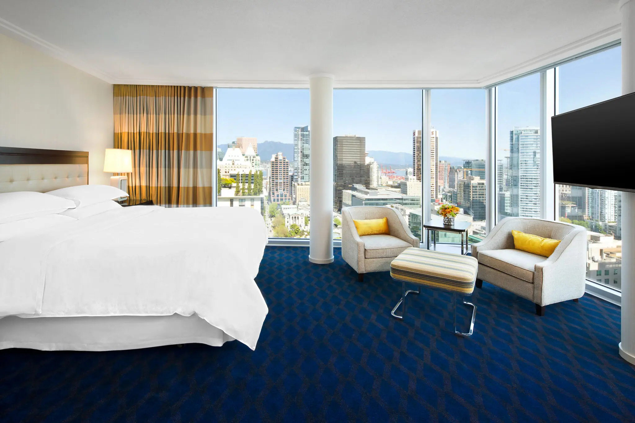 A bright, modern hotel room at the Sheraton Vancouver Wall Centre featuring floor-to-ceiling windows with a panoramic view of the downtown Vancouver skyline and mountains. The room includes a plush king bed with white linens, a blue patterned carpet, and a seating area with two grey armchairs, yellow accent pillows, and an ottoman.