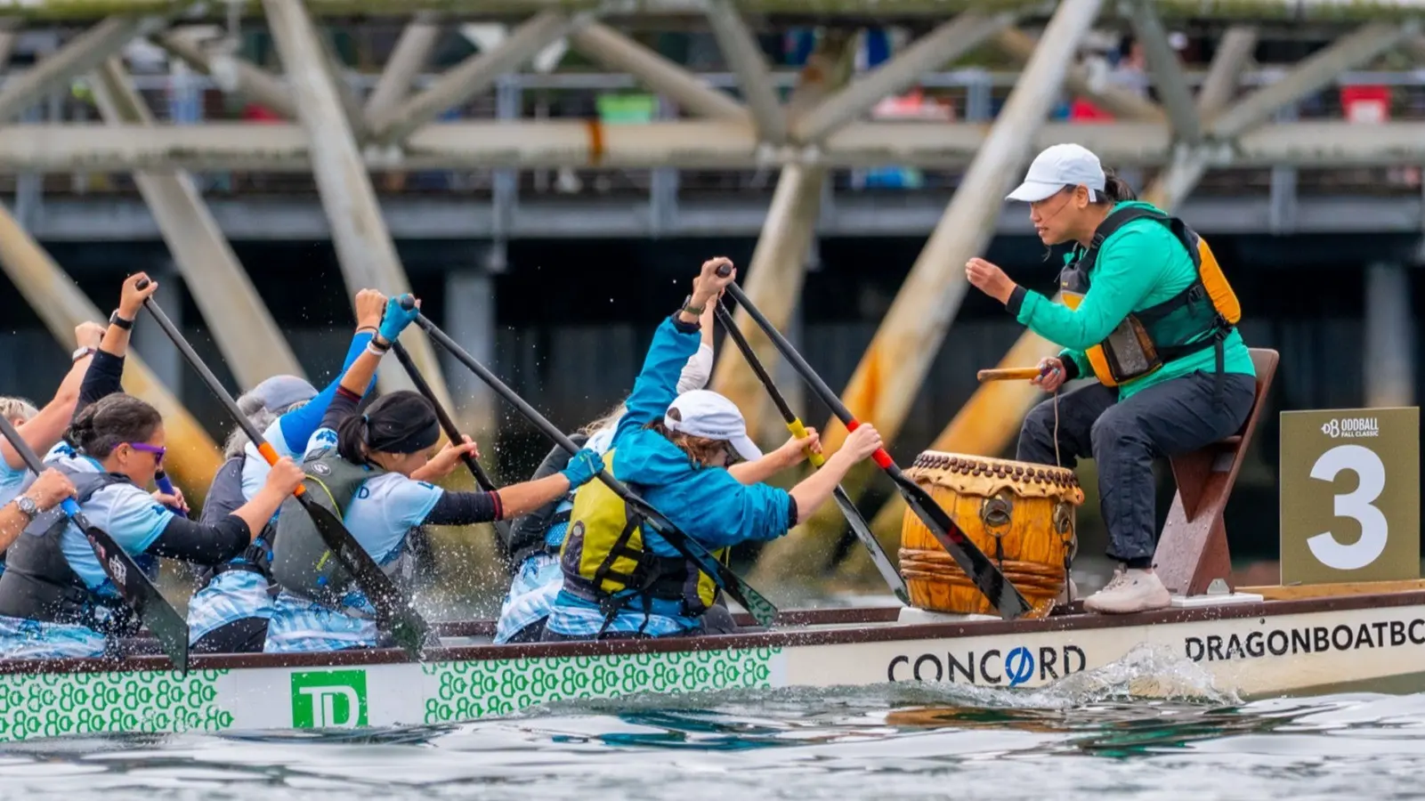 A team paddling a dragon boat with a drummer in a green jacket beating a drum at the front, near a sign with the number 3.