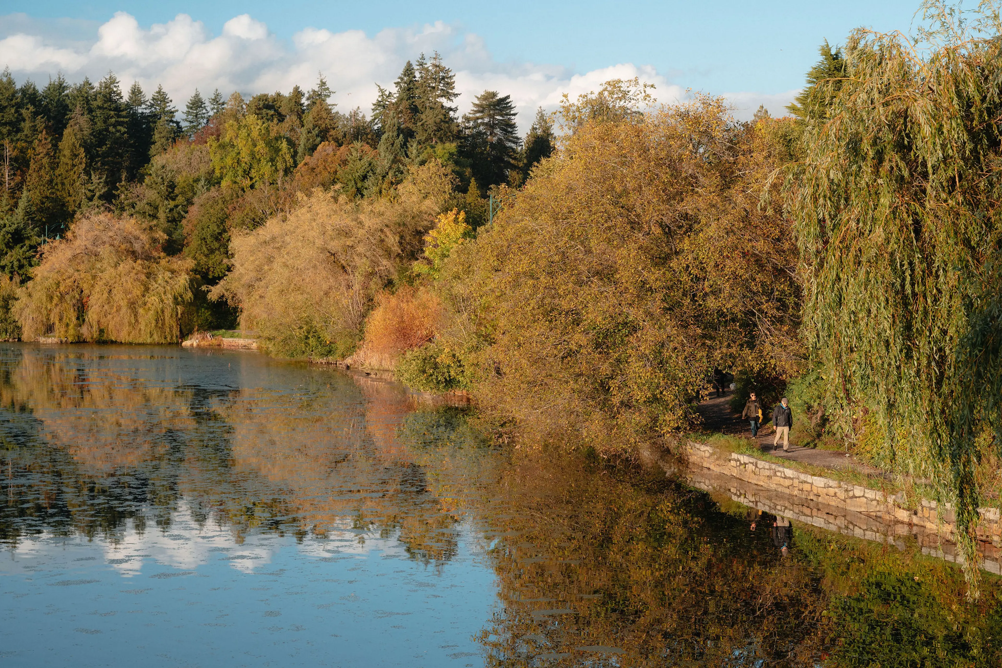 Two people walking on a path beside a calm lake with autumn trees reflecting in the water under a blue sky.