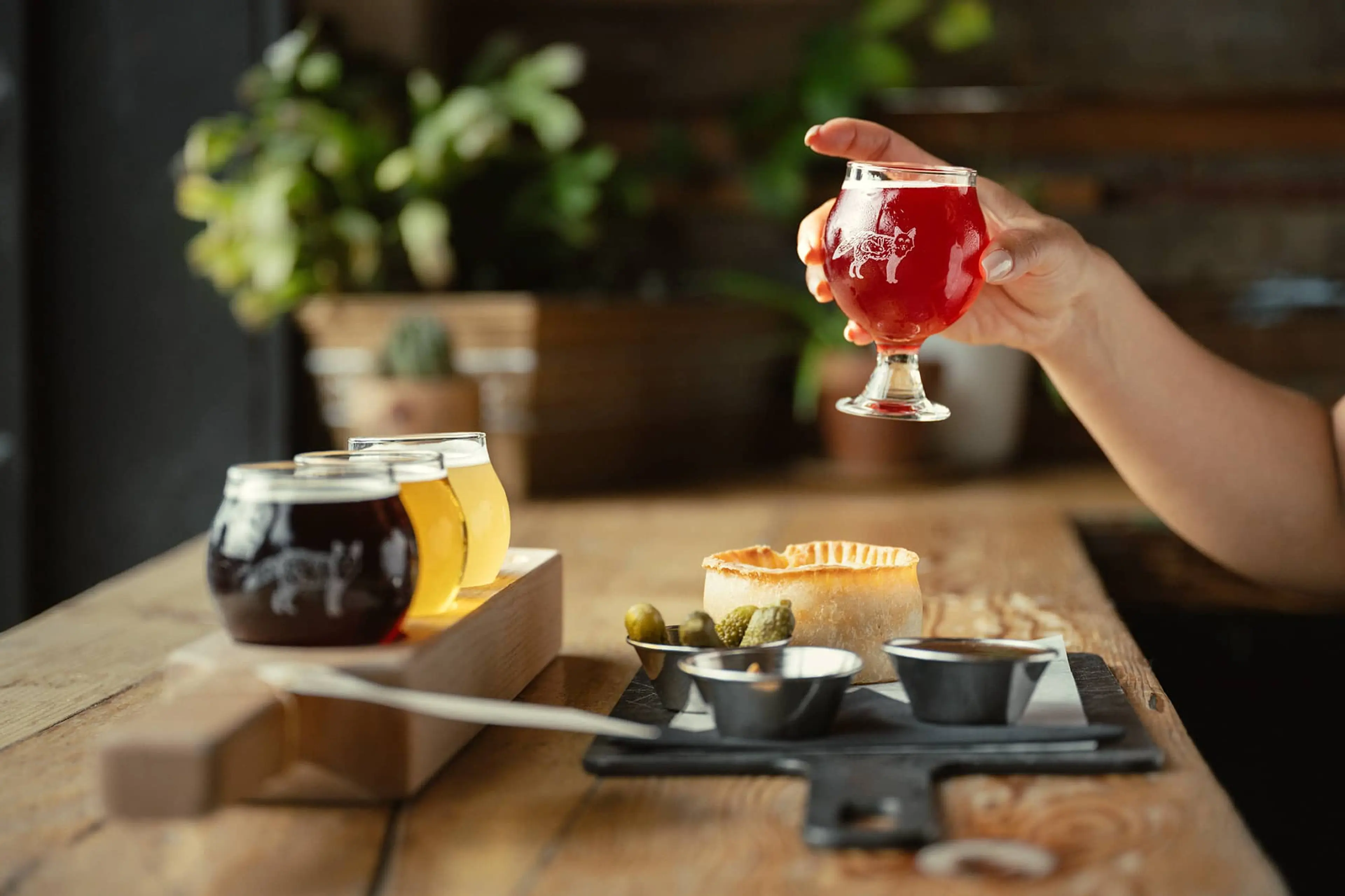A hand holding a glass of red beer above a wooden table with a flight of three beers, a small pie, pickles, and dipping sauces.