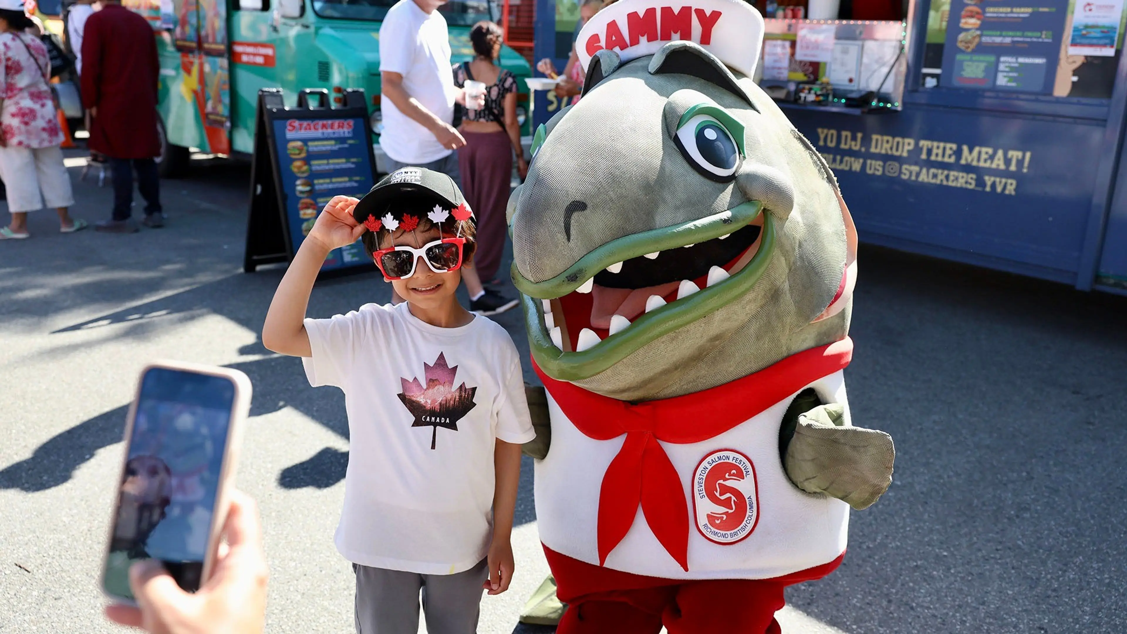 A child wearing a Canada-themed shirt, sunglasses, and a cap poses next to a person in a large fish mascot costume named Sammy at an outdoor event.
