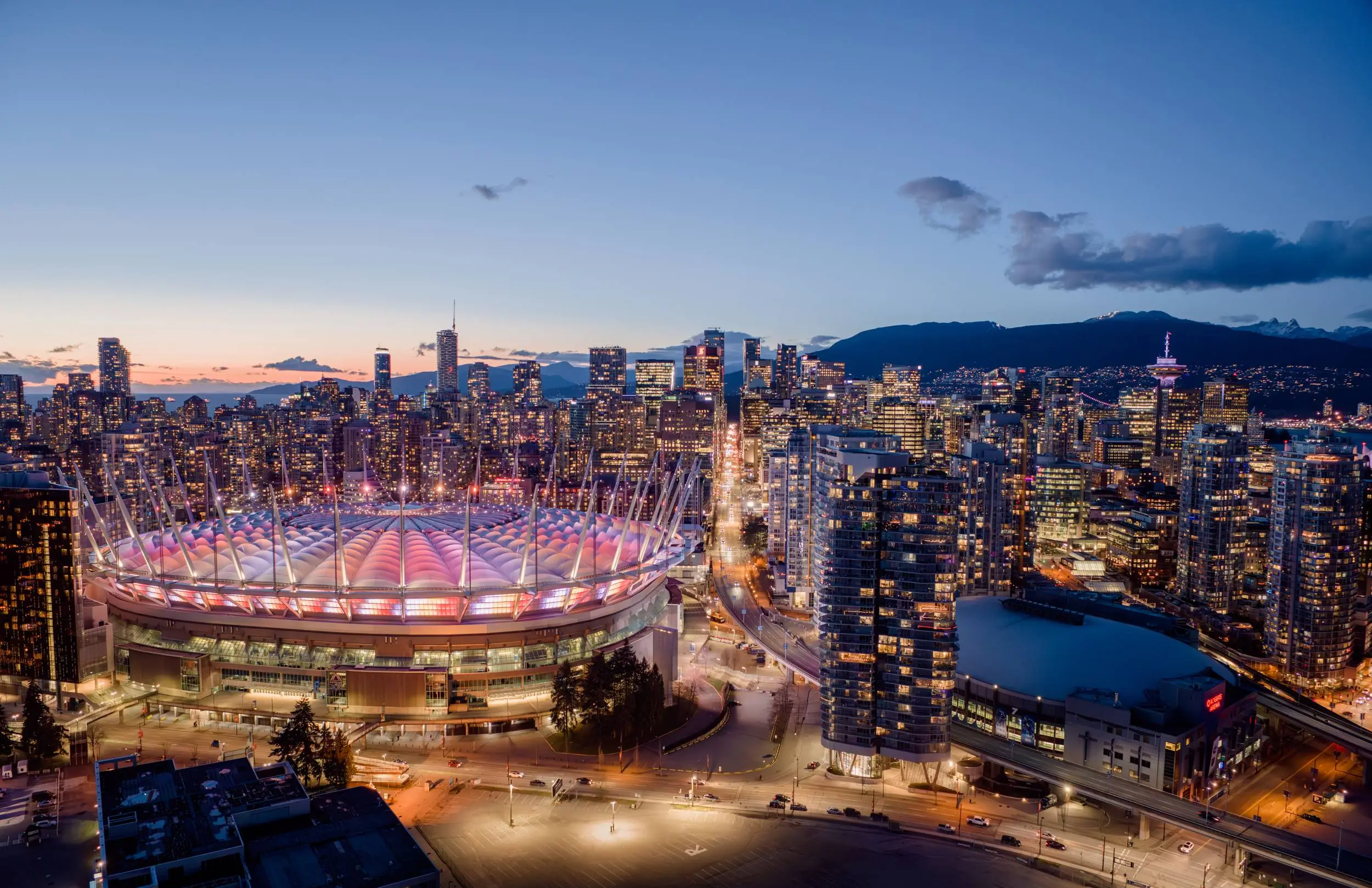 City skyline at dusk with a large stadium lit in pink and purple lights in the foreground and mountains in the background.