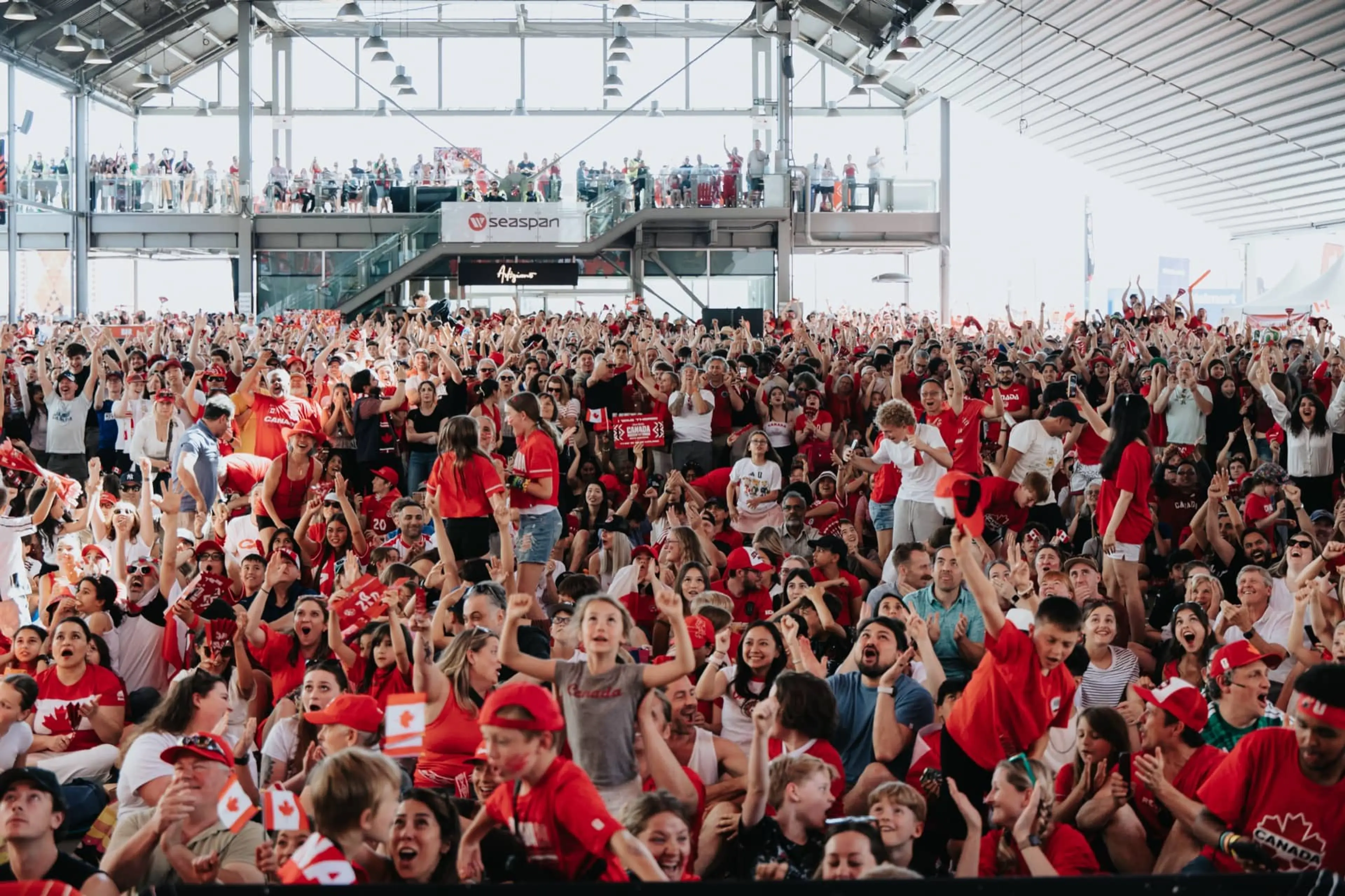 Large crowd of people in a stadium wearing red and white, many holding Canadian flags and cheering.