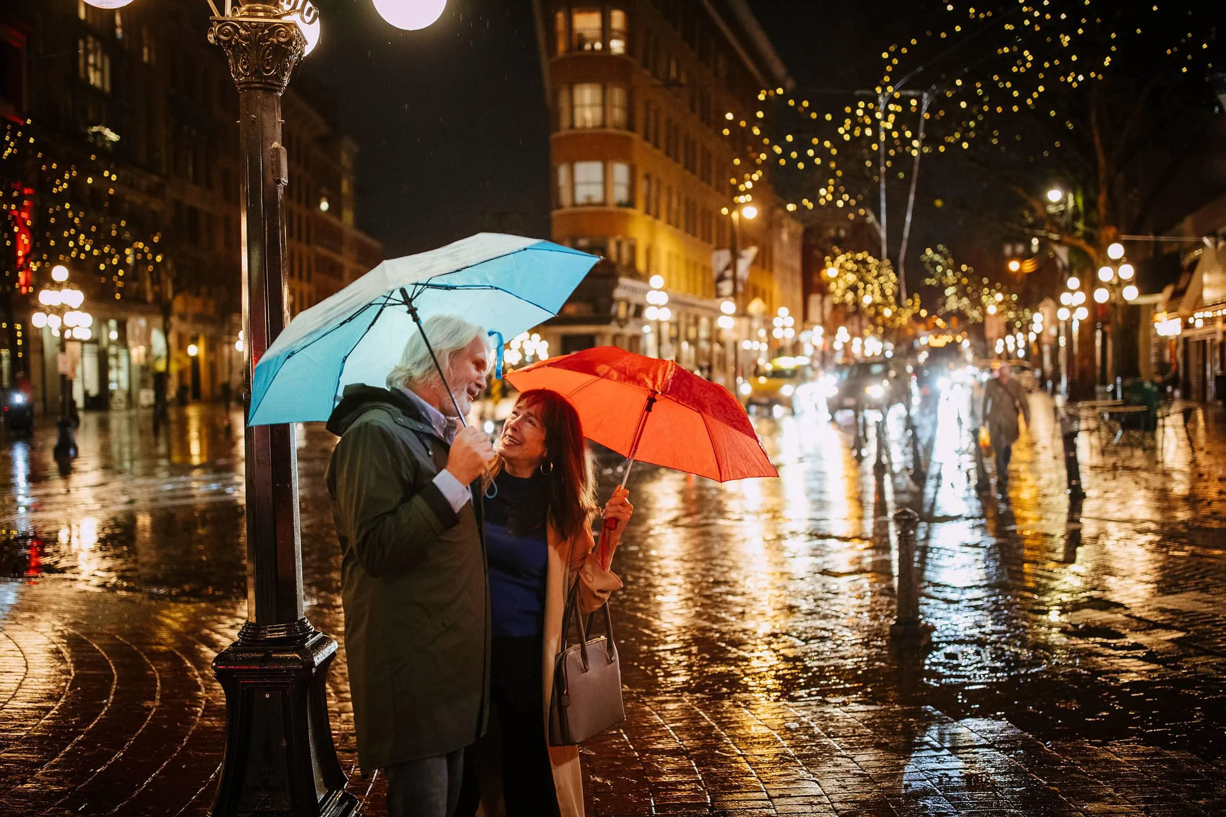 Older couple standing under umbrellas on a wet city street at night with festive lights and reflections on the pavement.