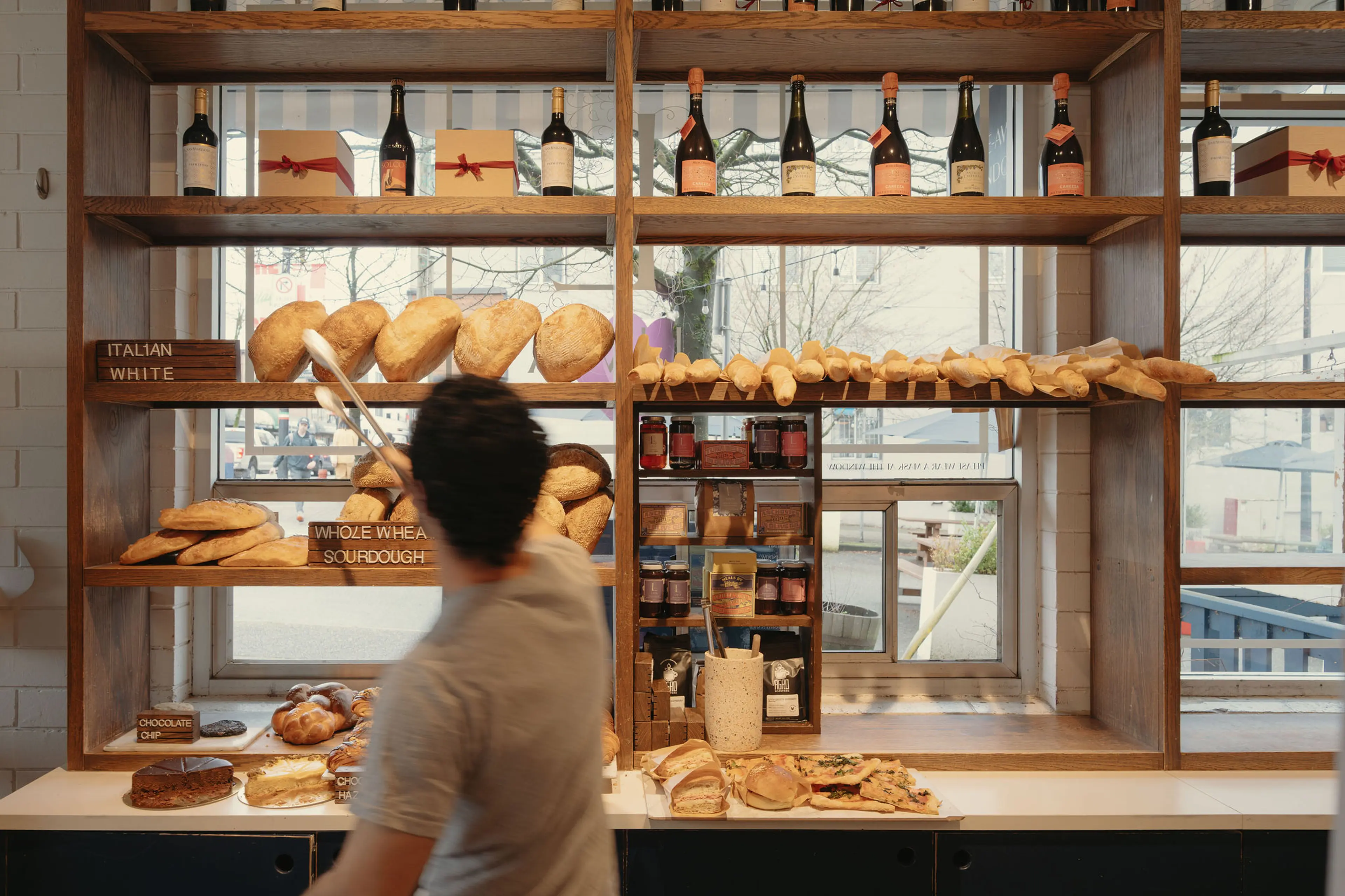 Person using tongs to pick bread from wooden shelves filled with various breads and bottles in a bakery.