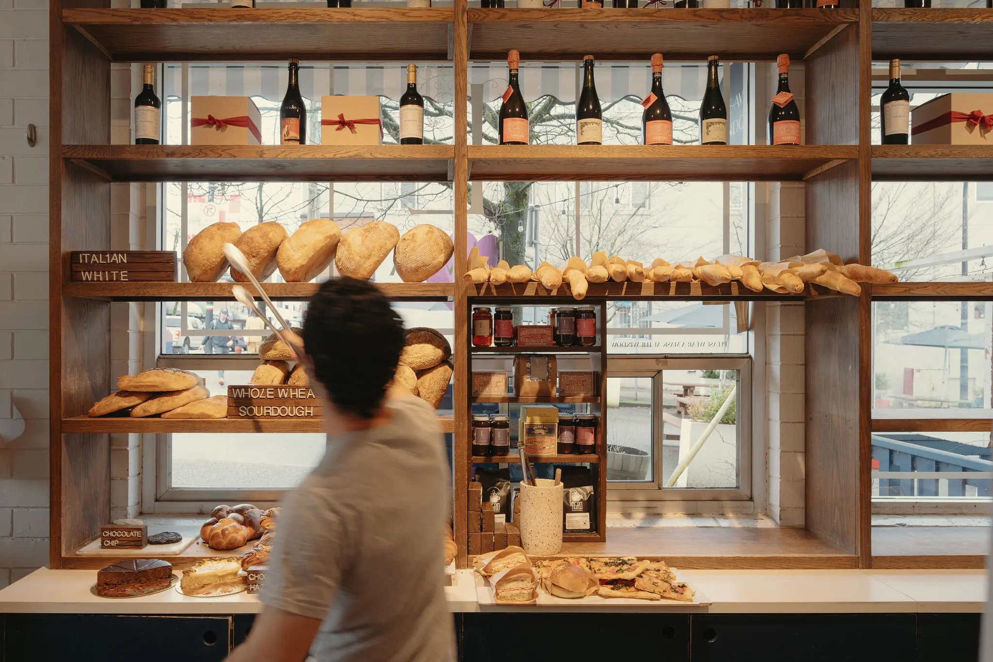 Person using tongs to pick bread from wooden shelves filled with various breads and bottles in a bakery.