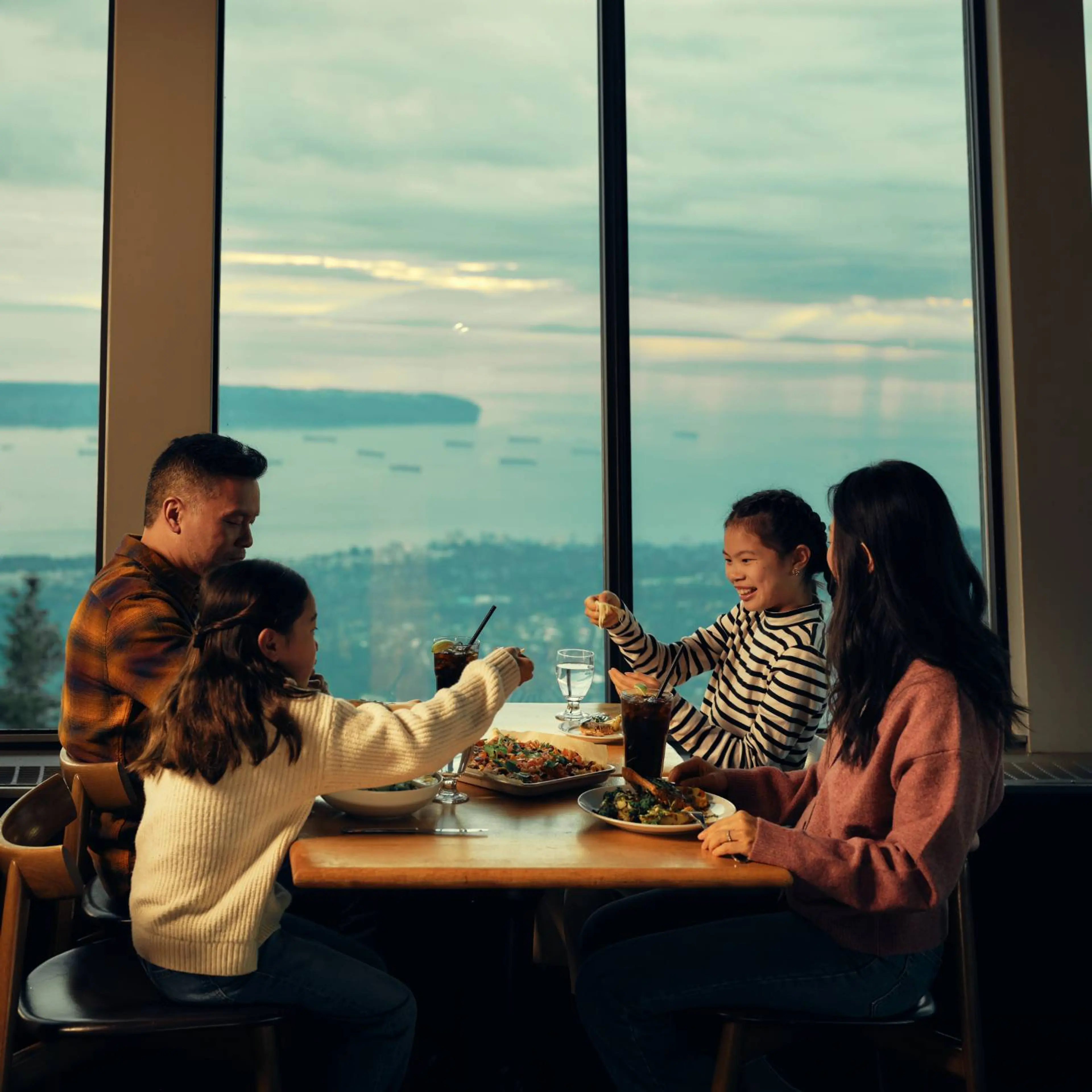 Family of four enjoying a meal together at a table by a large window with a scenic ocean view.