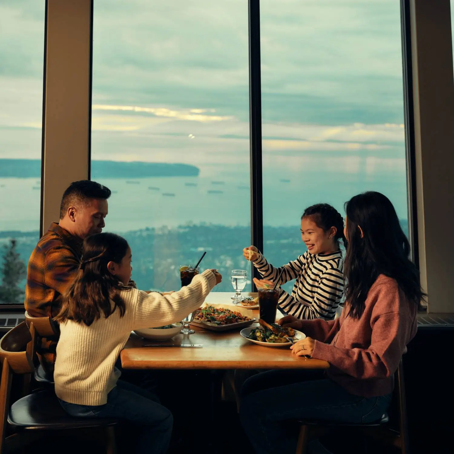 Family of four enjoying a meal together at a table by a large window with a scenic ocean view.