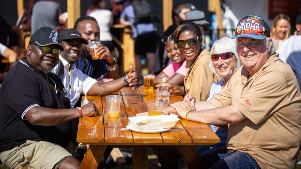 Seven people sitting at a wooden picnic table outdoors, smiling and enjoying drinks in plastic cups.