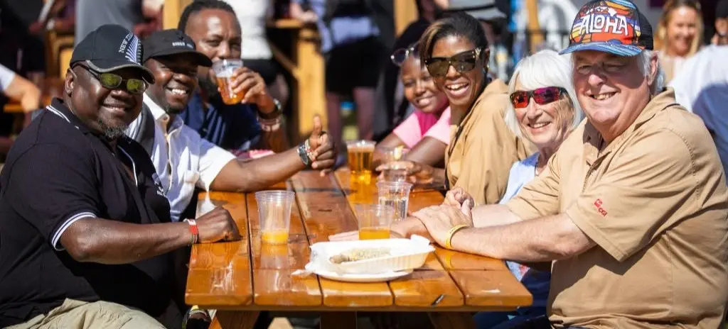 Seven people sitting at a wooden picnic table outdoors, smiling and enjoying drinks in plastic cups.