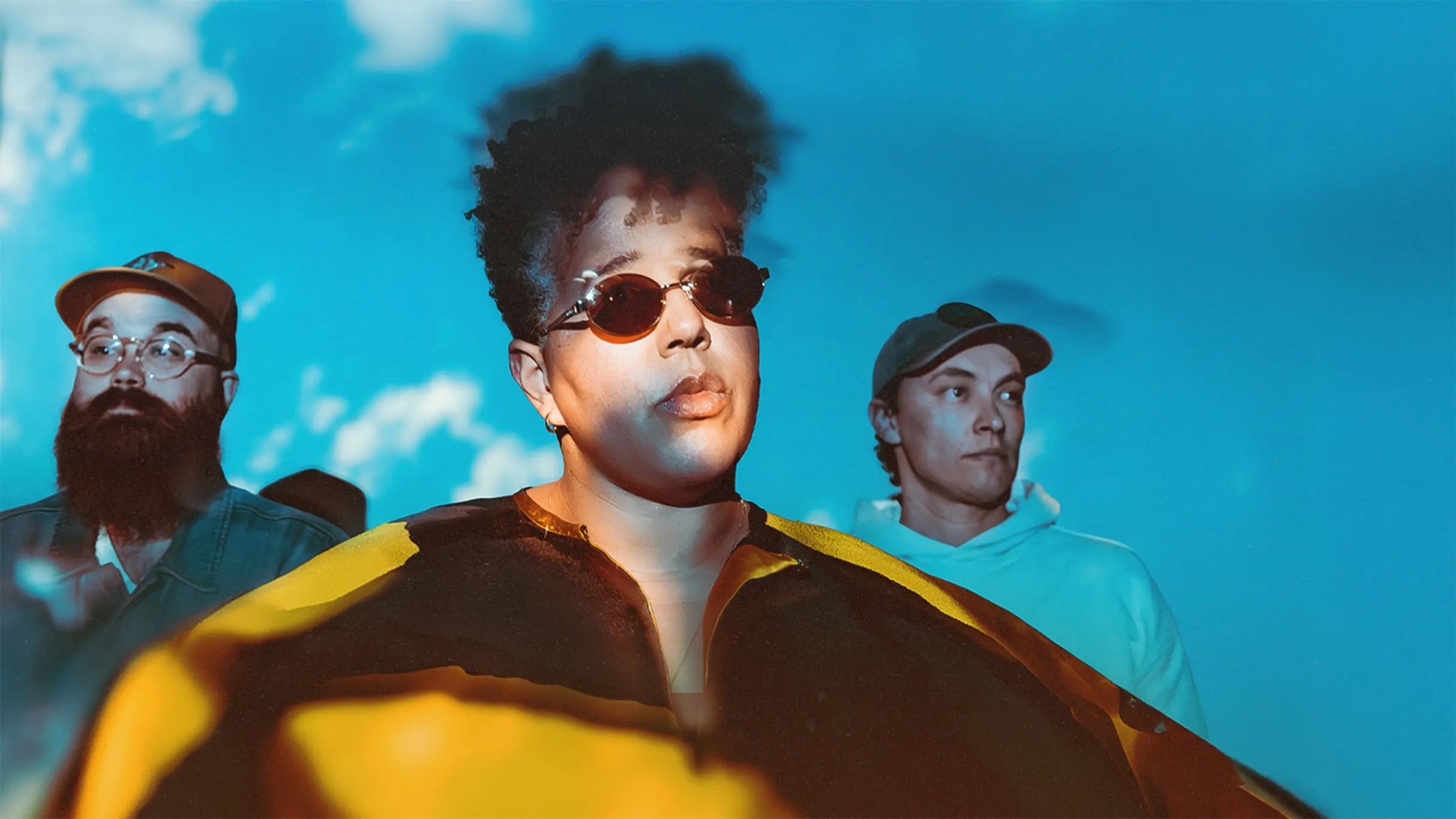 Three people standing against a blue sky background, one wearing sunglasses and a yellow and black jacket, another with a beard and glasses, and the third in a light hoodie and cap.