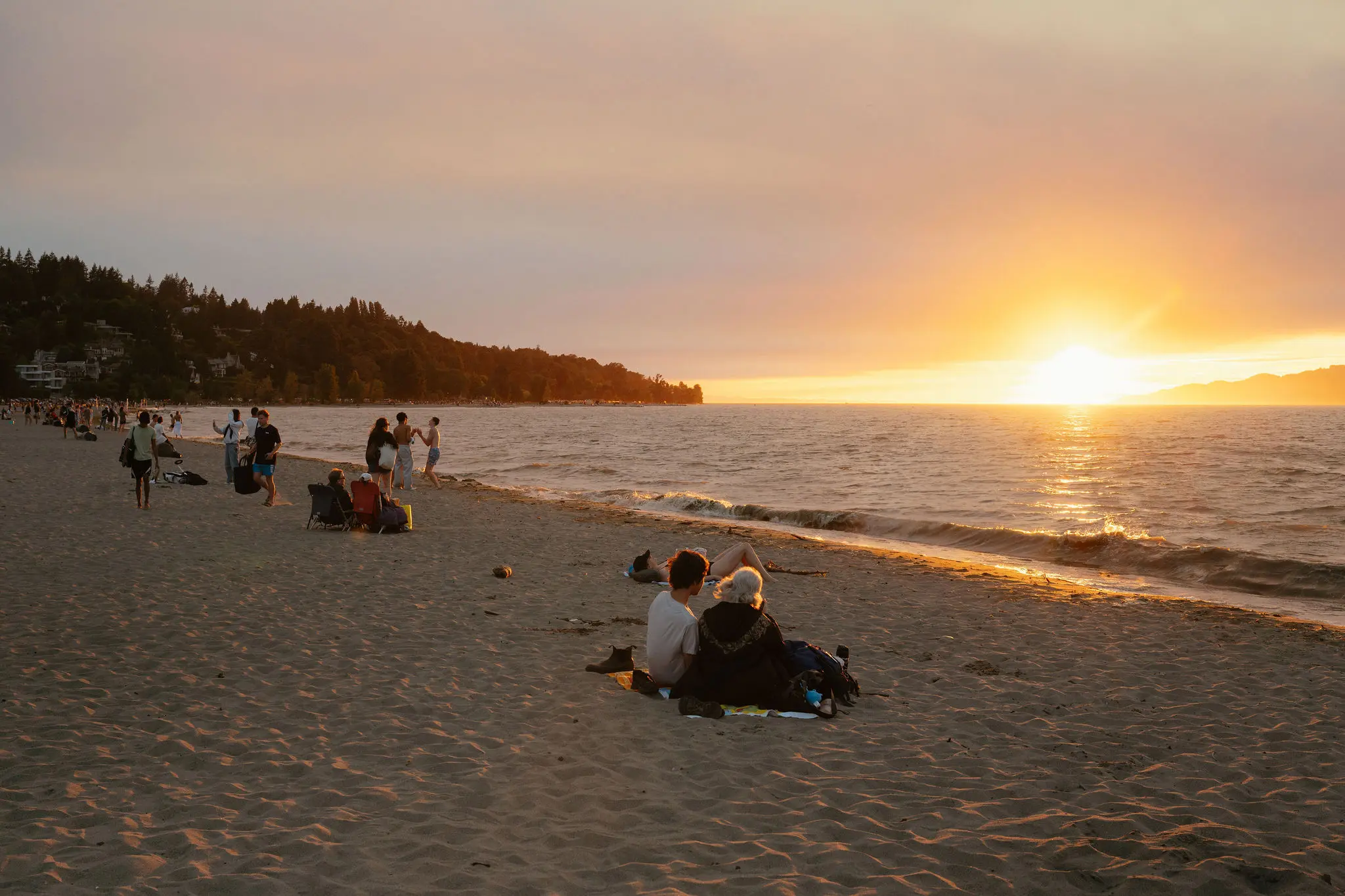People sitting and walking on a sandy beach at sunset with the sun low over the water.
