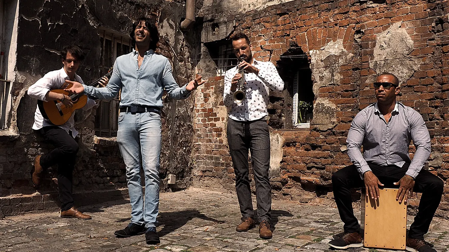Four people performing music outdoors against a weathered brick wall; one playing guitar, one singing, one playing saxophone, and one playing a wooden box drum.
