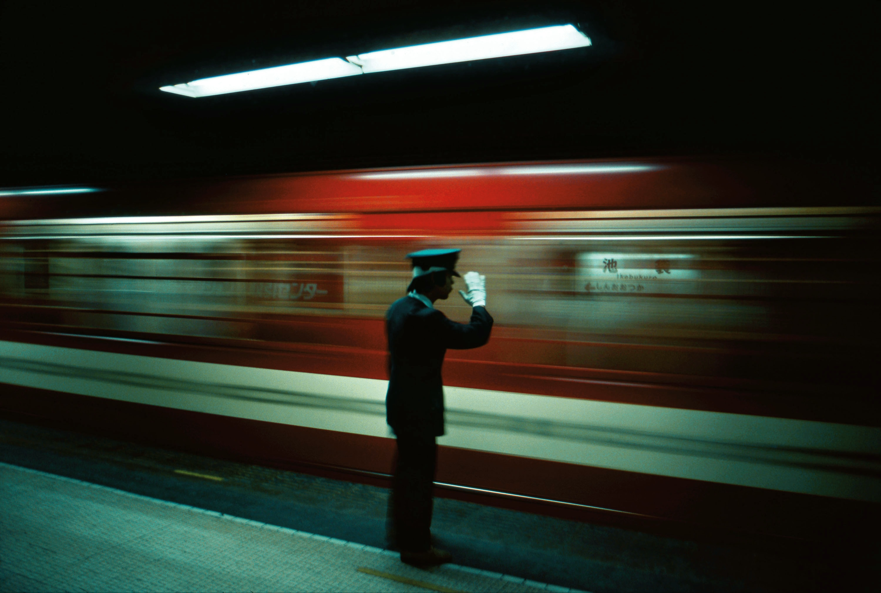 A person in uniform and white gloves saluting on a subway platform with a red train passing by in motion blur.