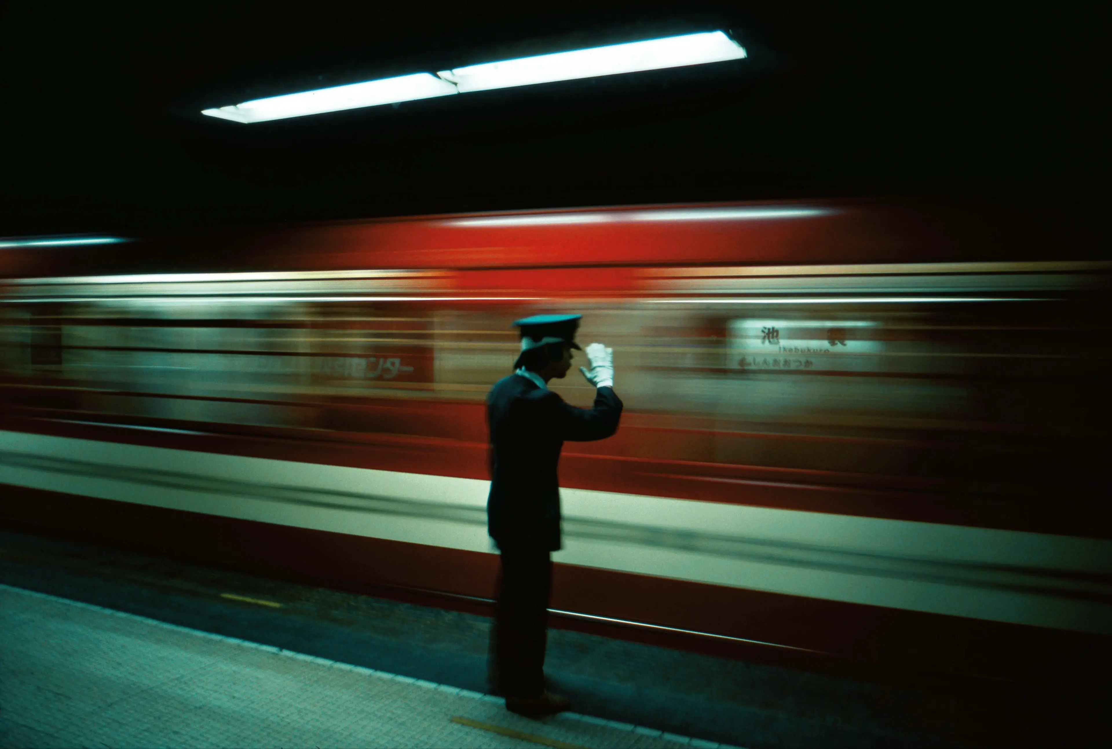 A person in uniform and white gloves saluting on a subway platform with a red train passing by in motion blur.