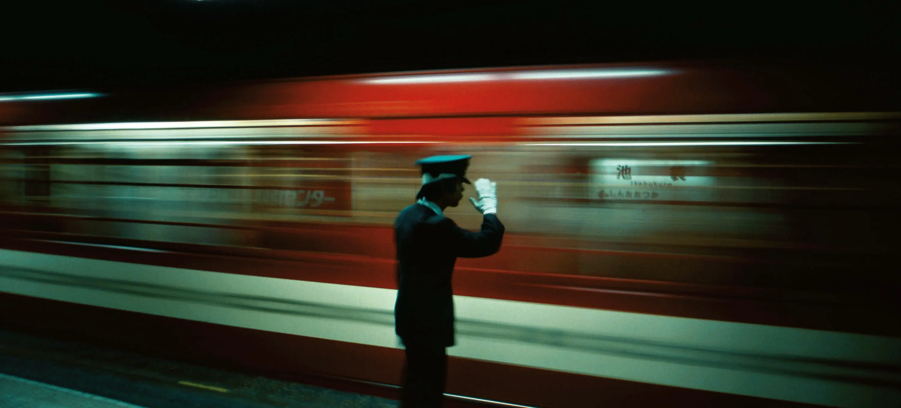 A person in uniform and white gloves saluting on a subway platform with a red train passing by in motion blur.