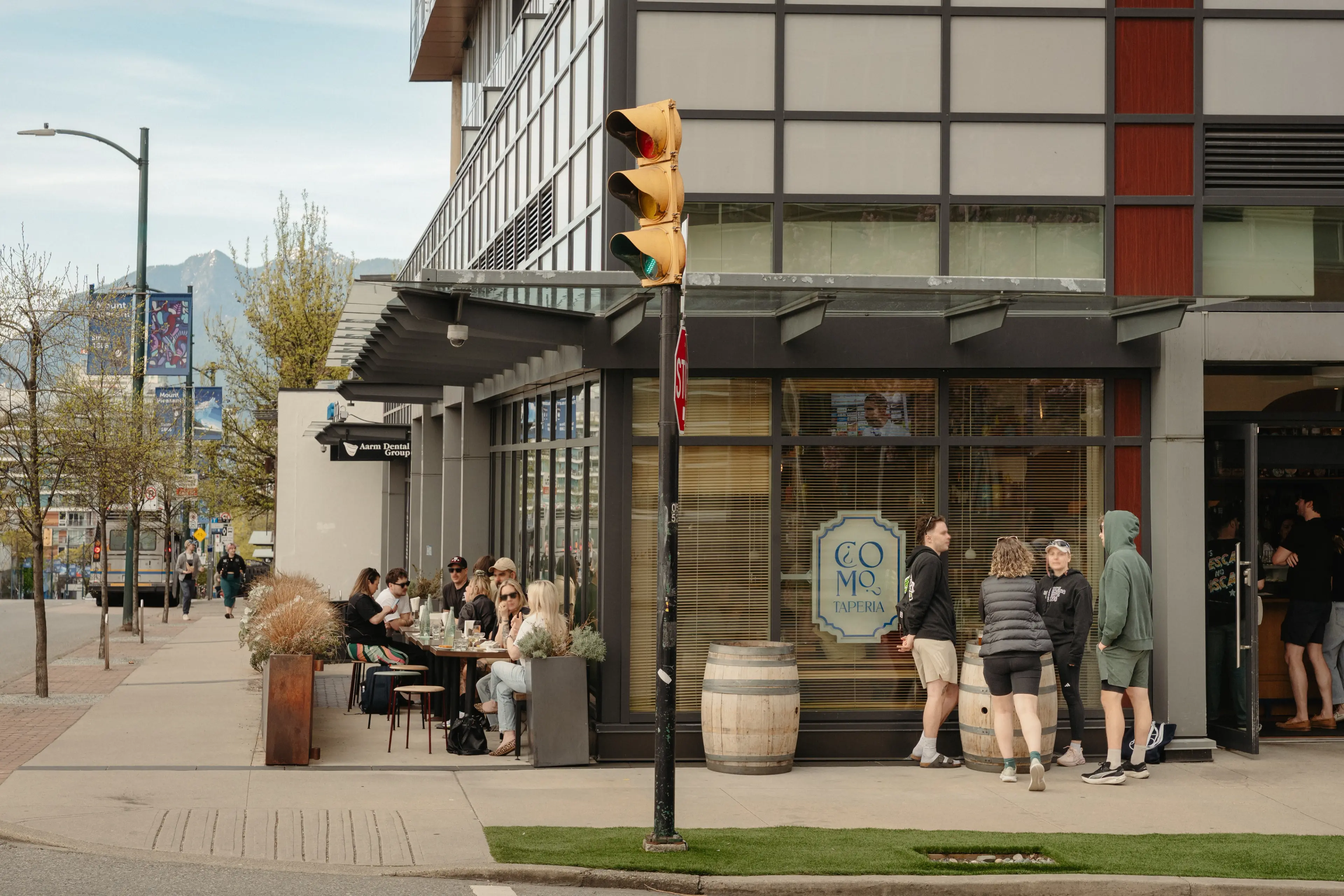 People sitting at outdoor tables and standing outside Como Taperia restaurant on a city sidewalk near a traffic light.
