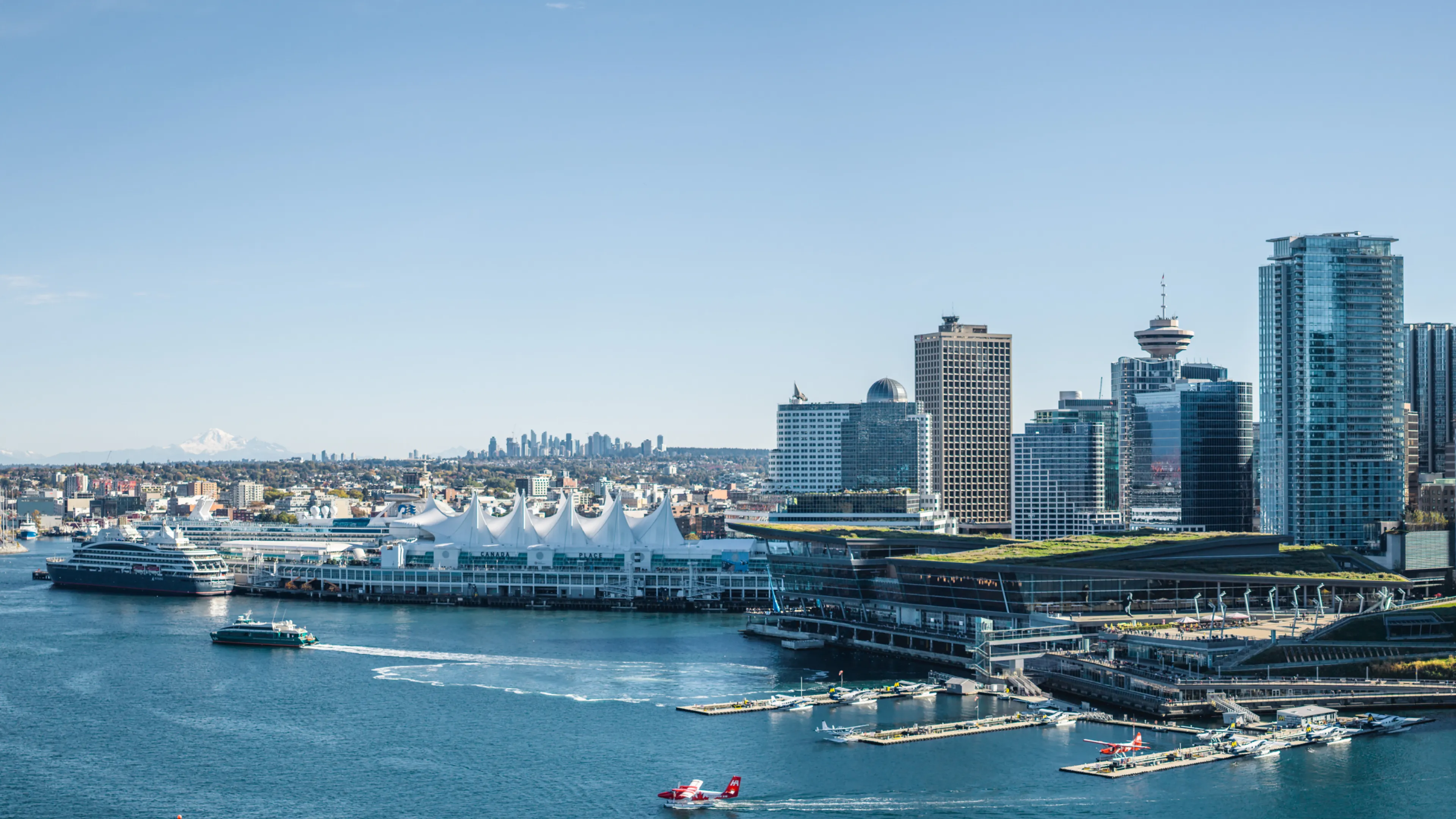 City skyline with modern buildings, waterfront with cruise ships, ferries, and seaplanes on a clear day.