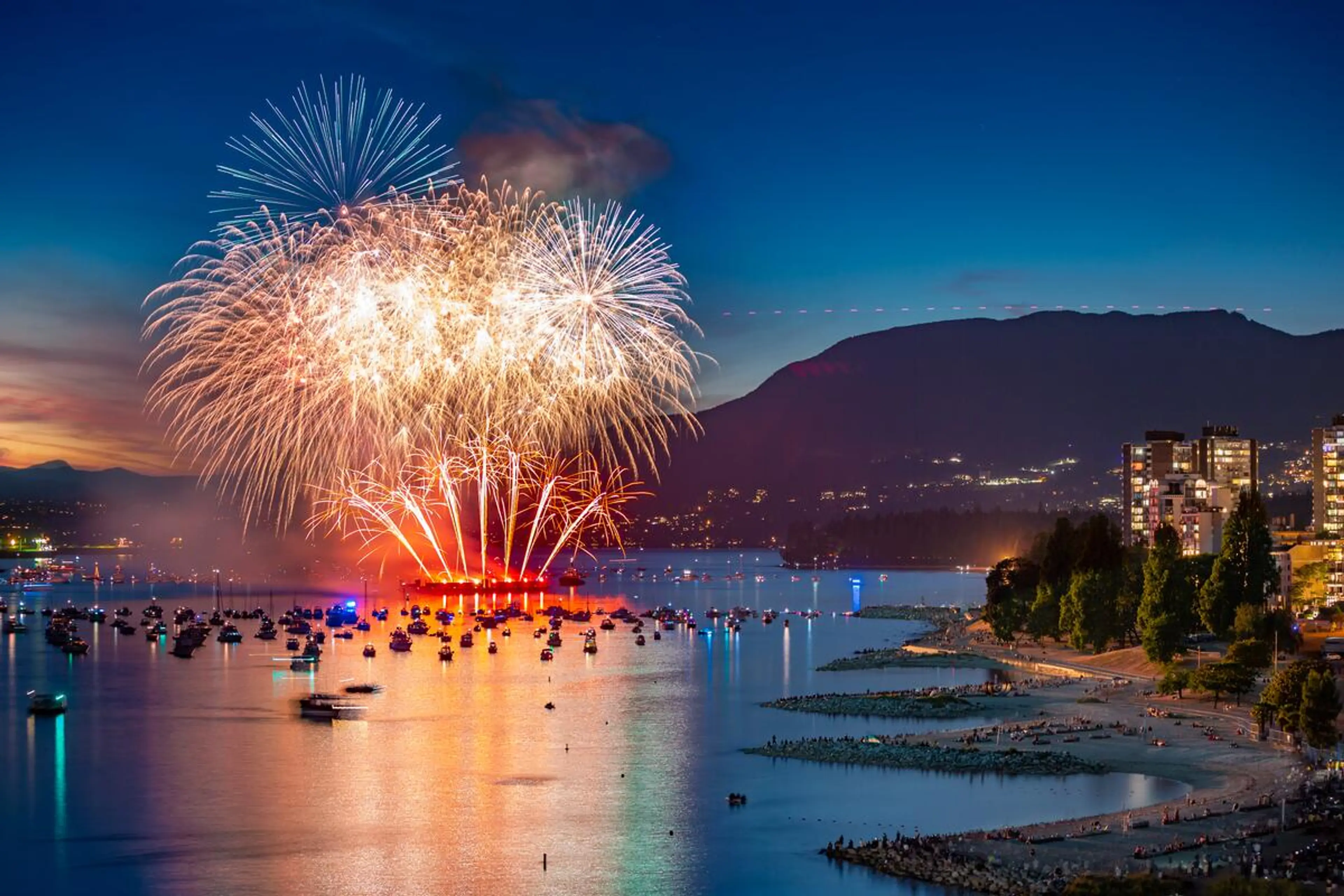 Fireworks exploding over a harbor filled with boats at dusk, with a cityscape and mountains in the background.