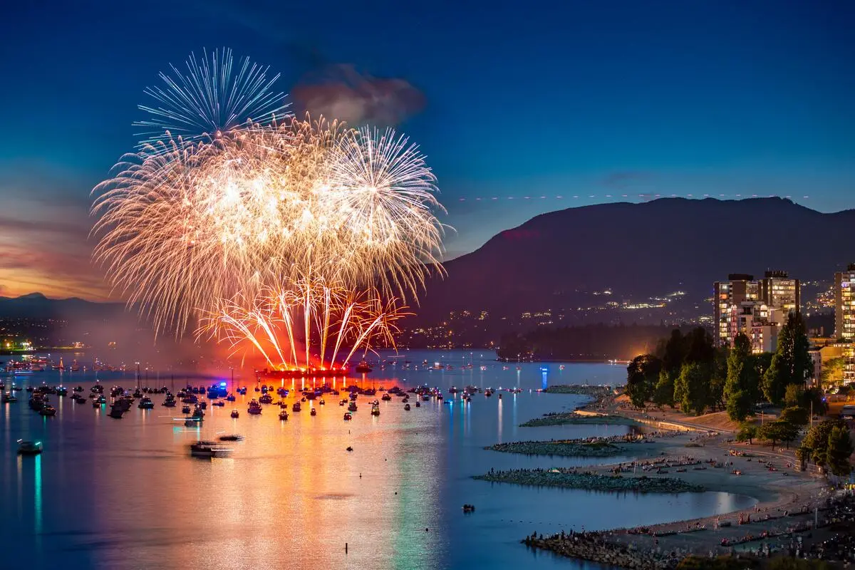 Fireworks exploding over a harbor filled with boats at dusk, with a cityscape and mountains in the background.