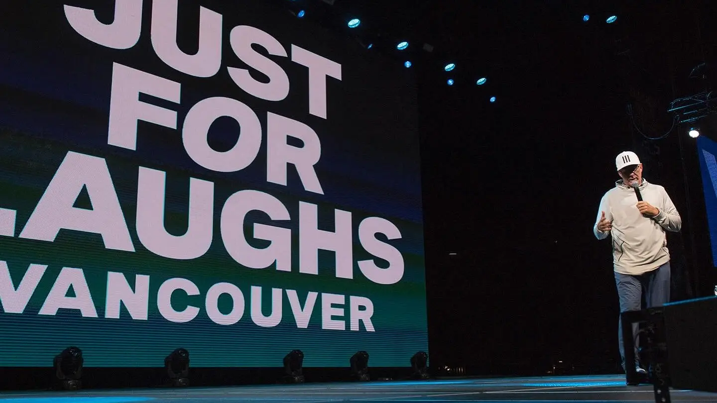 Comedian on stage holding a microphone with a large screen behind displaying JUST FOR LAUGHS VANCOUVER.